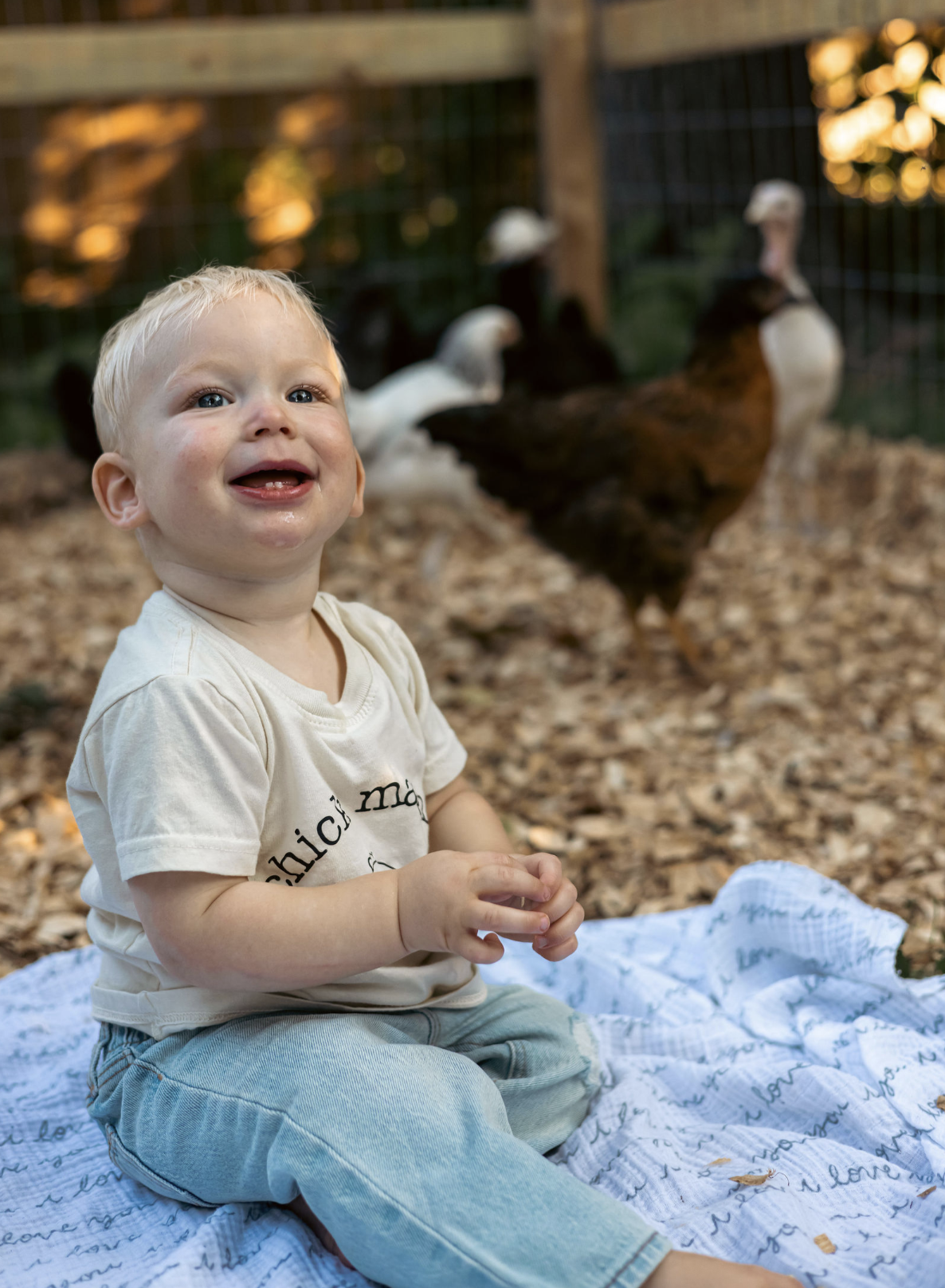 A young child with blonde hair and blue eyes sitting on a blanket outdoors, smiling with a few teeth visible, near a chicken coop with chickens in the background.