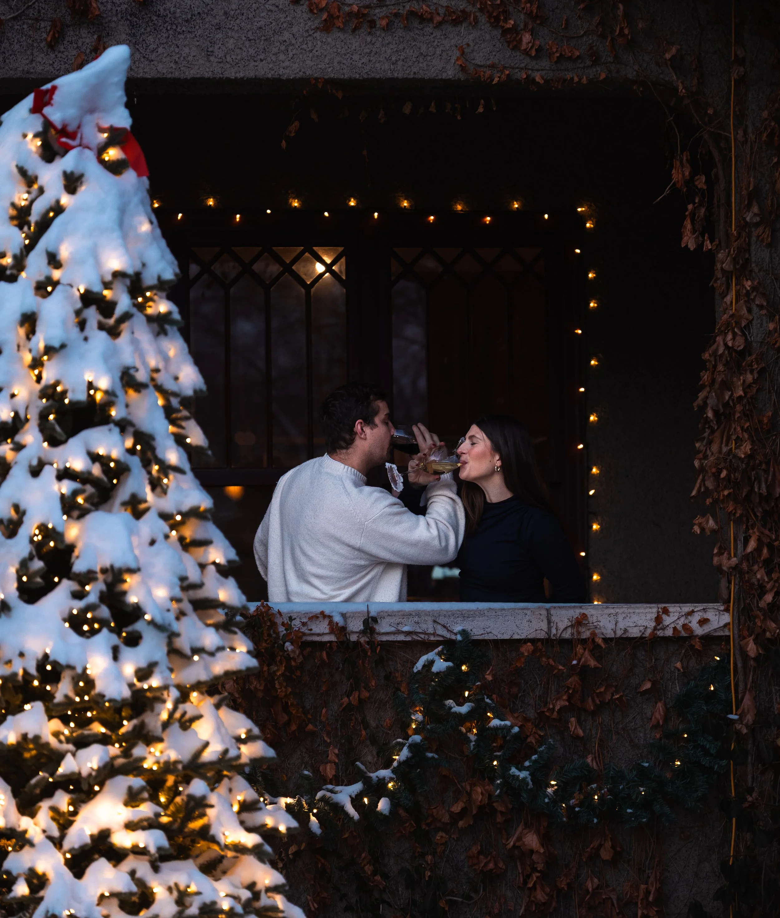 A couple is celebrating Christmas outdoors with a decorated, snow-covered Christmas tree and string lights, sharing a toast.