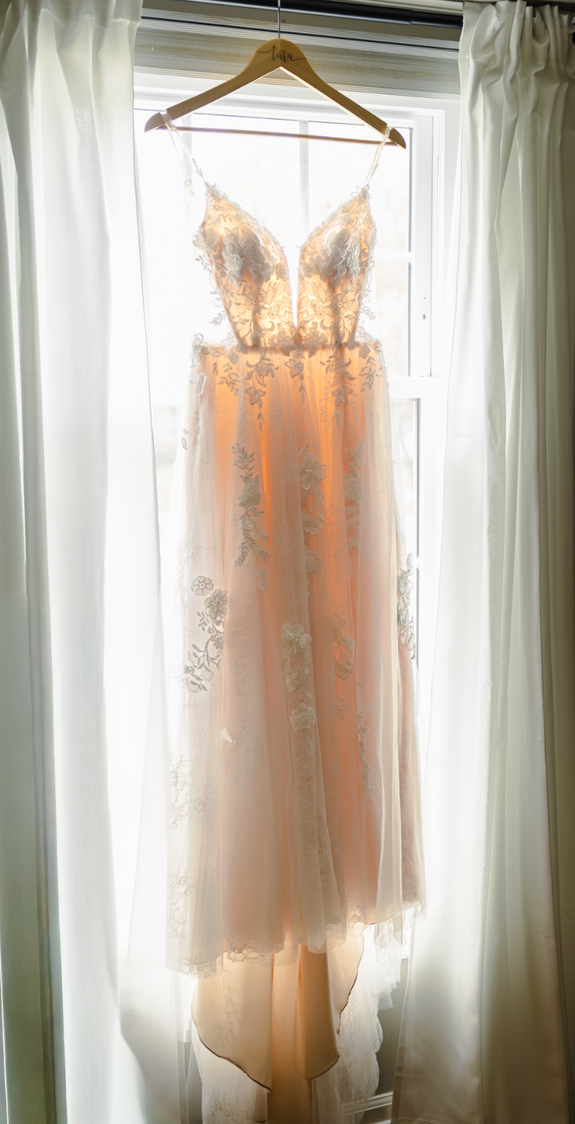 Wedding dress hanging on a hanger in front of a window with curtains, backlit by sunlight.