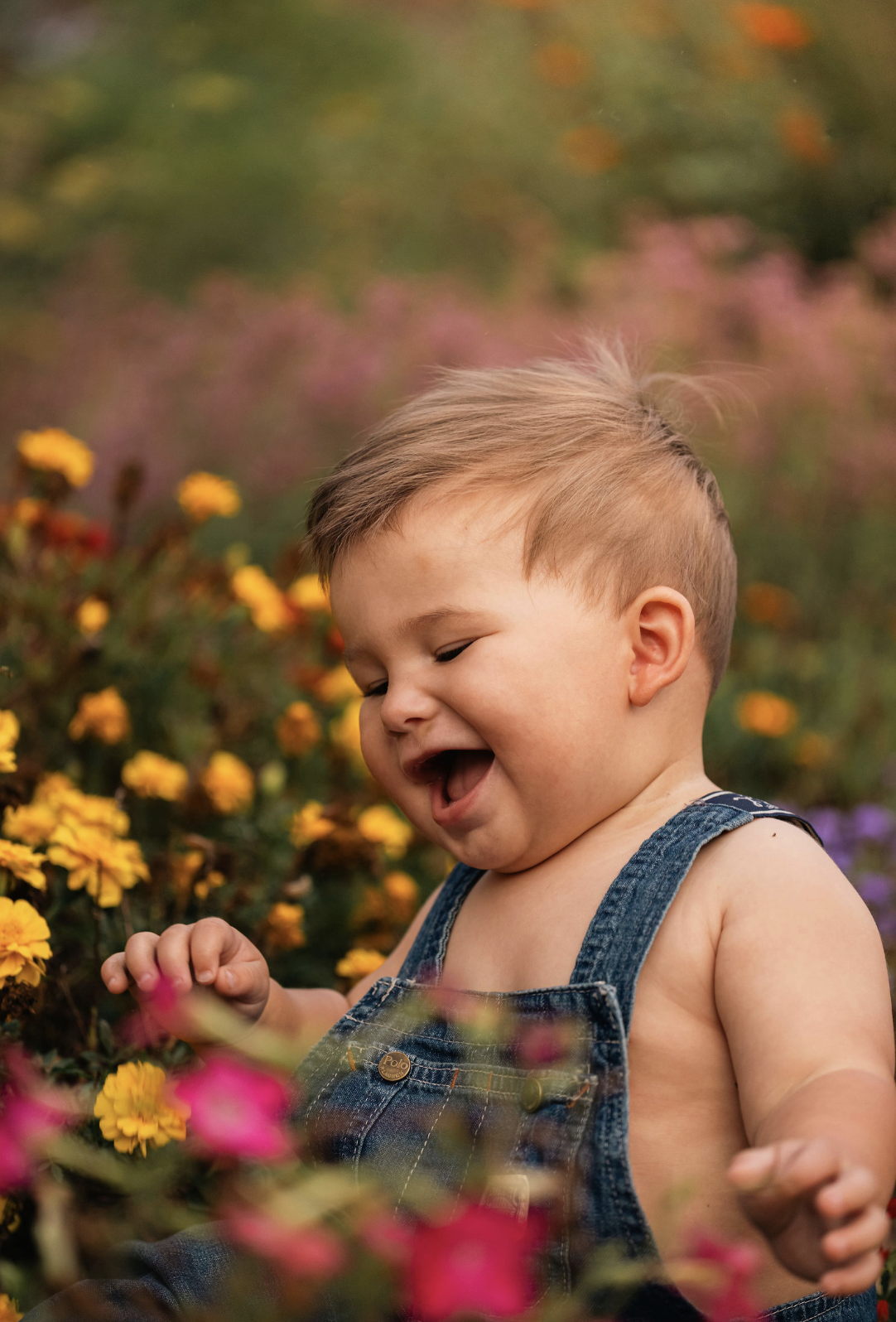 A young child with short hair, wearing denim overalls, smiling and laughing among colorful flowers in a garden.