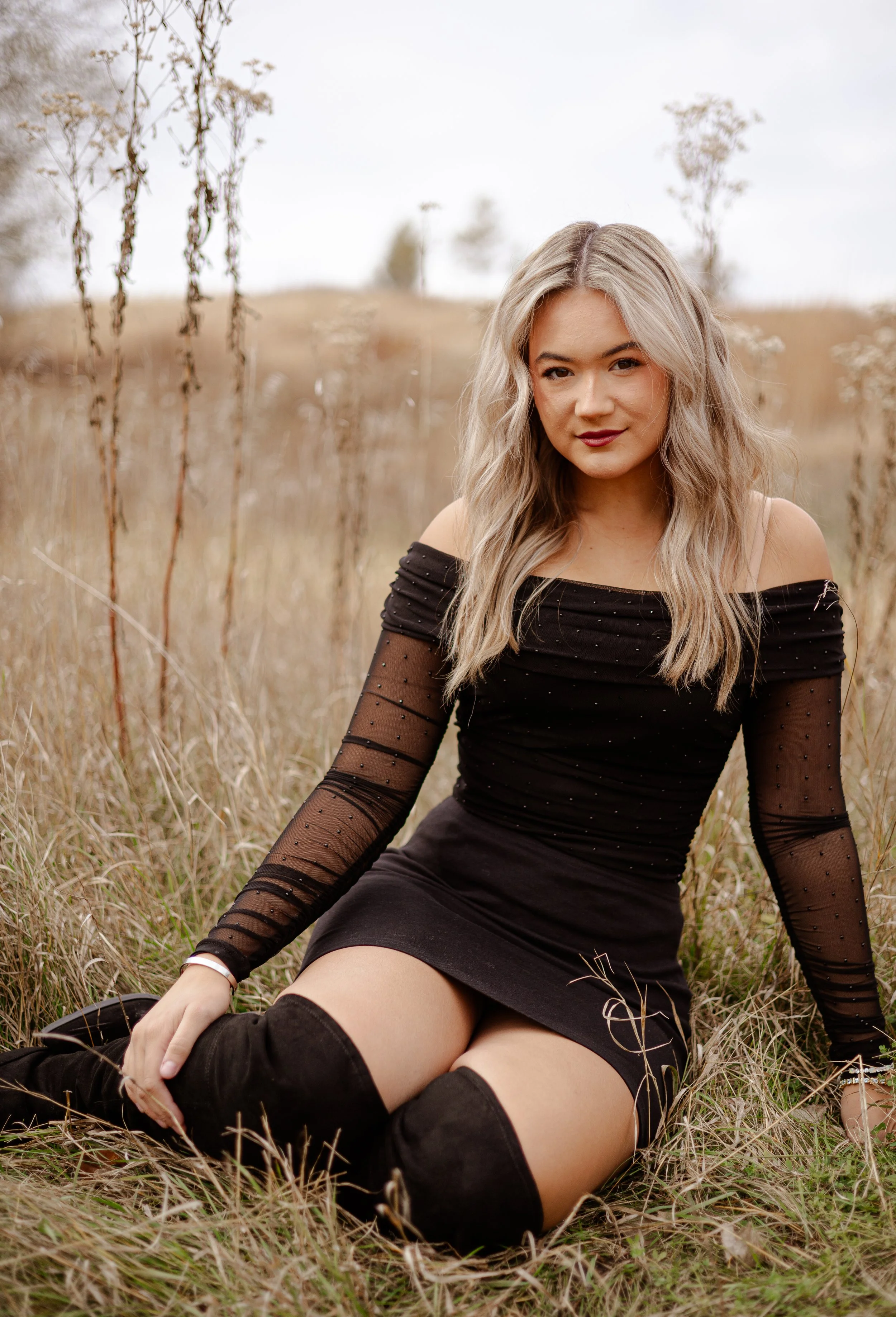 A young woman sitting in a field of tall, dry grass with a few trees in the background, wearing a black off-shoulder dress and black thigh-high boots.