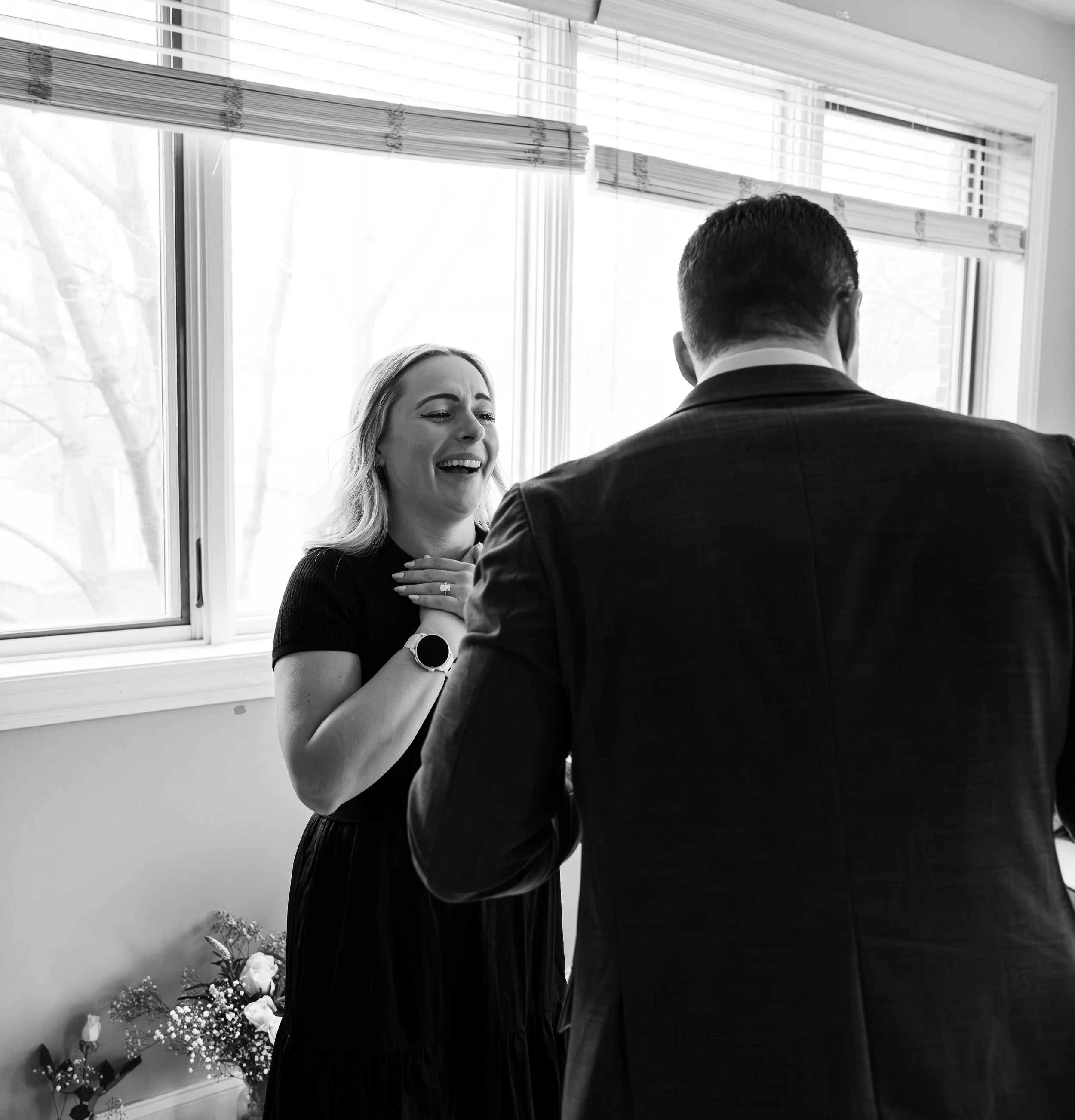 A woman with long hair and a man in a suit sharing a joyful moment near a window. The woman is smiling with her eyes closed, and they appear to be happy. There are flowers and plants in the background.