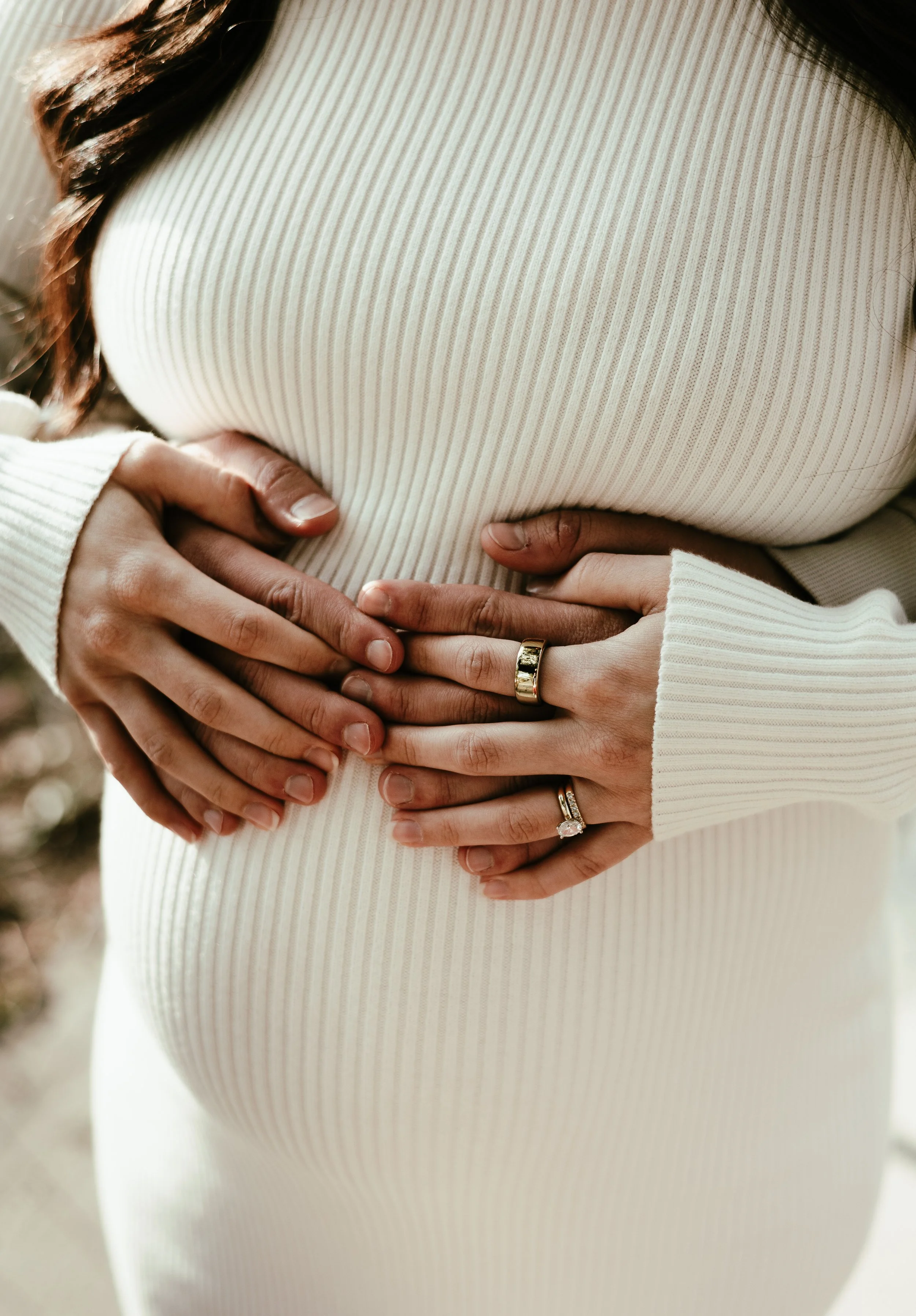 Close-up of multiple hands resting on a pregnant woman's belly, with rings visible on some fingers, wearing a cream-colored ribbed top.