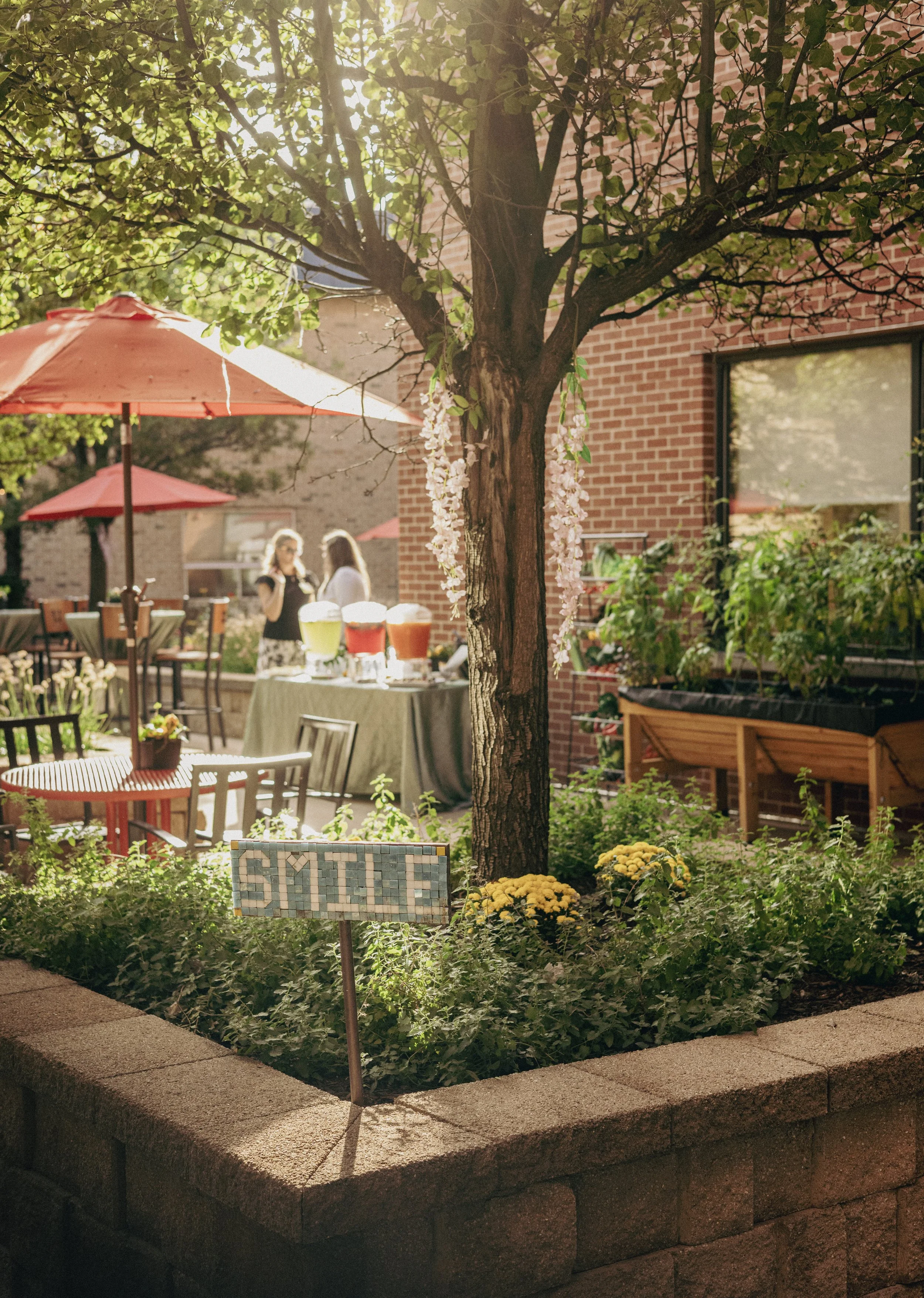 A garden with a brick building in the background, a tree with hanging flowers, outdoor tables and chairs, umbrellas, and a sign that reads 'SMILE' surrounded by plants and yellow flowers.