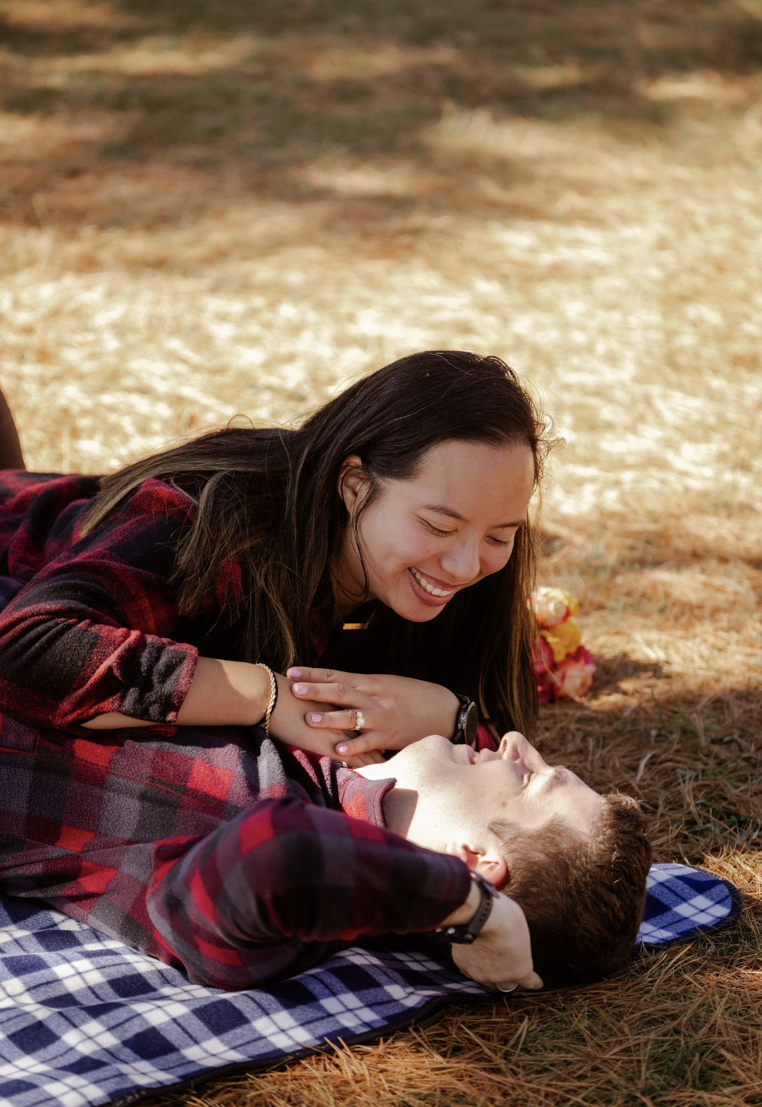 A woman and a man lying on a checkered blanket in a pine forest, smiling and looking at each other.