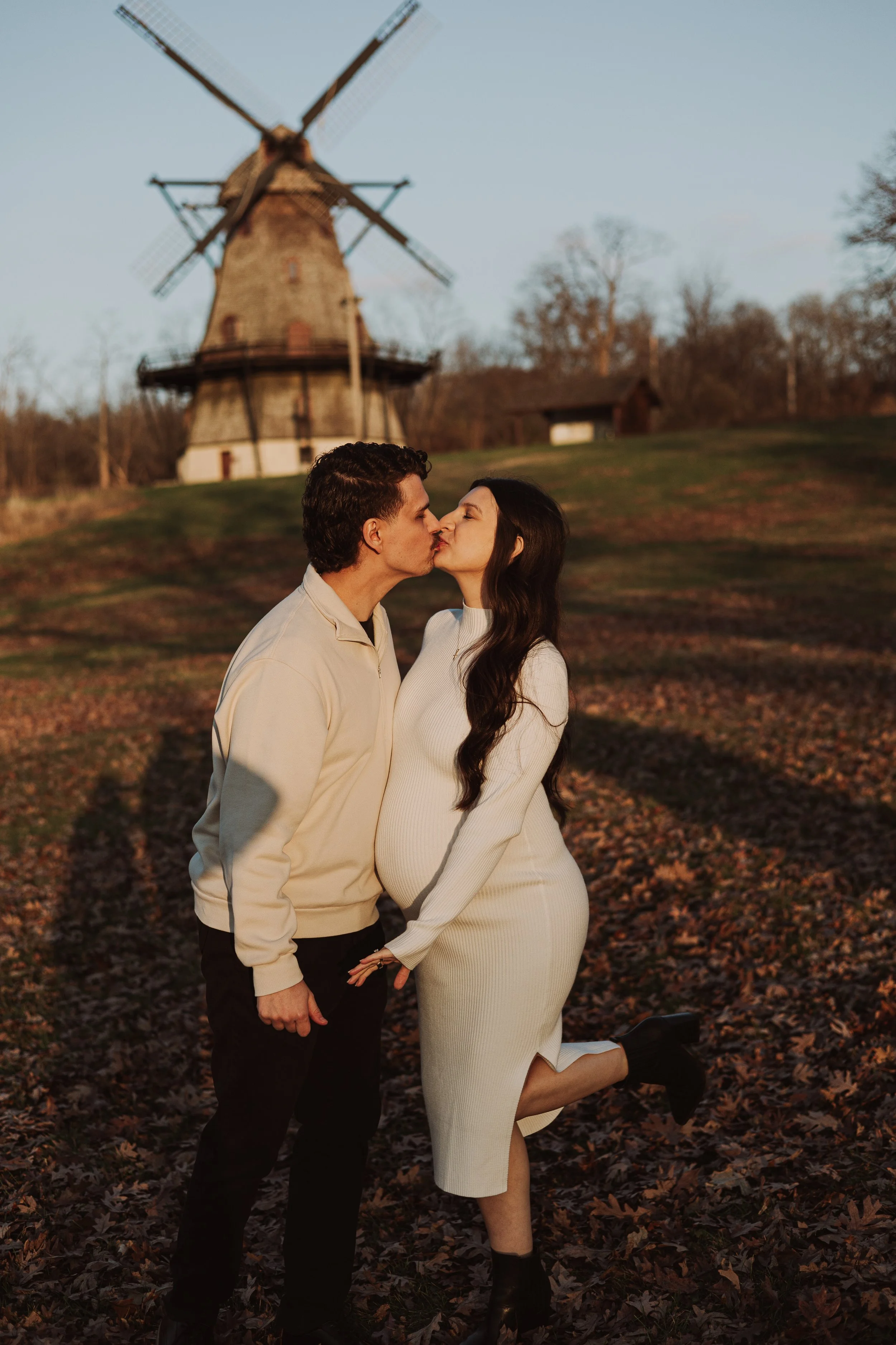 A man and woman kissing outdoors during autumn, standing on a leaf-covered ground with a traditional windmill in the background.