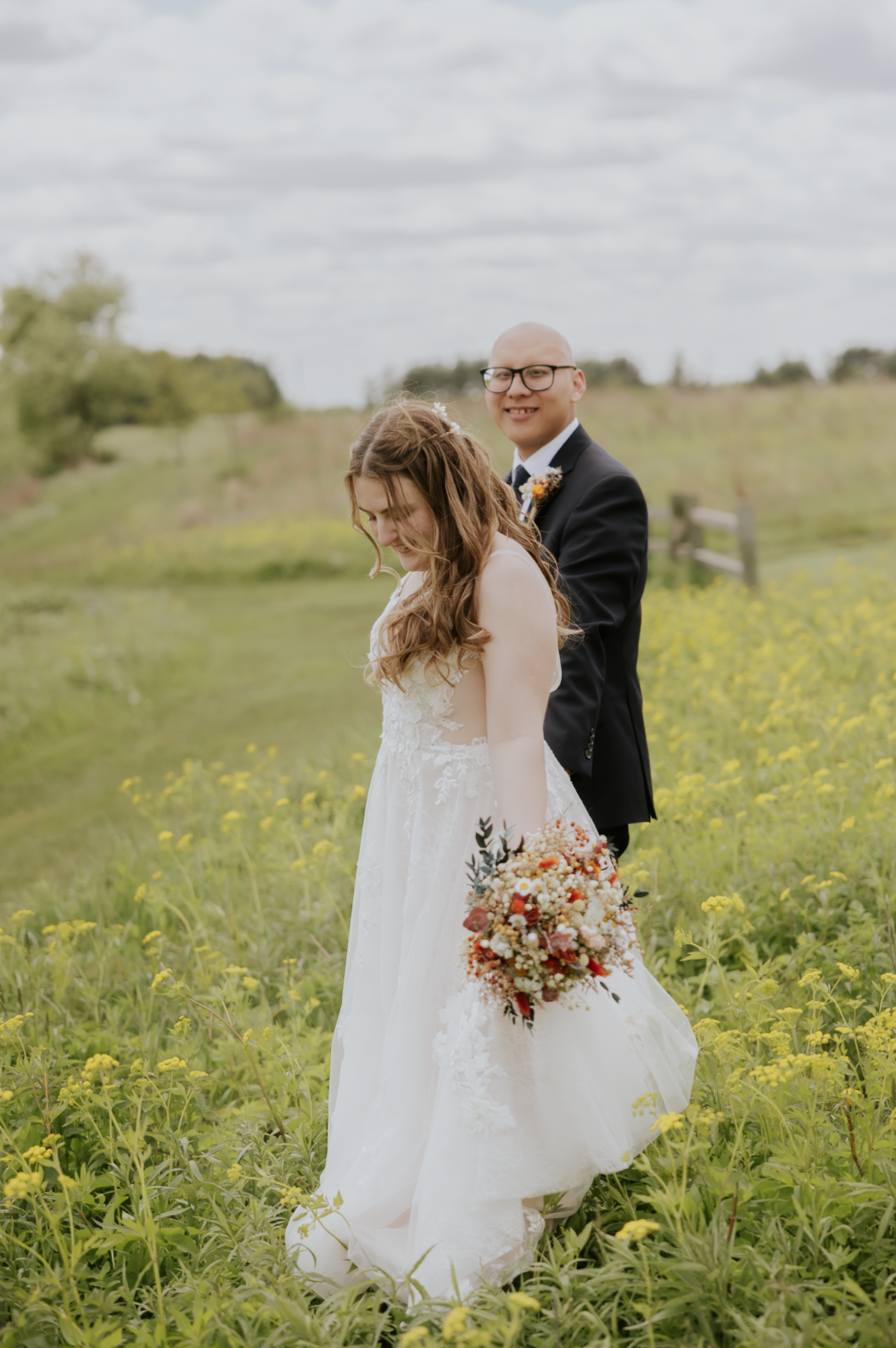 A bride in a white wedding dress holding a bouquet of flowers stands in a field of yellow flowers, with a groom in a dark suit smiling behind her, outdoors on a cloudy day.
