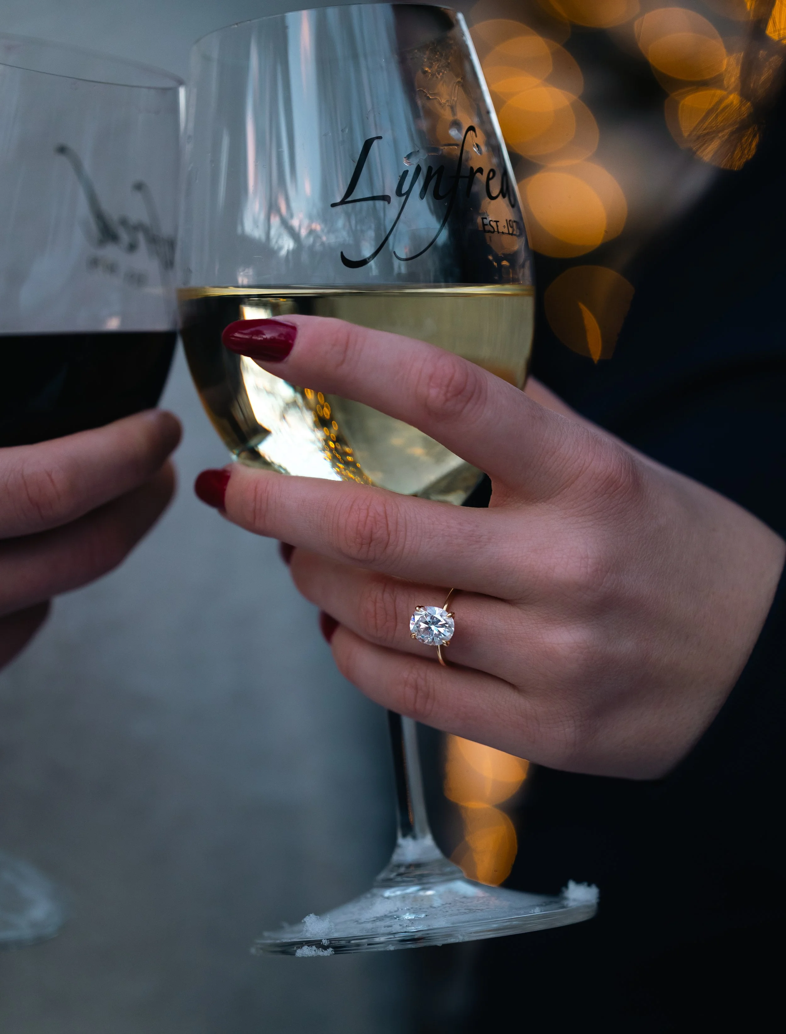 Close-up of two people clinking glasses of white wine, one person wearing a ring with a large diamond. The glasses have the word 'Lynred' printed on them. Background has warm bokeh lights.