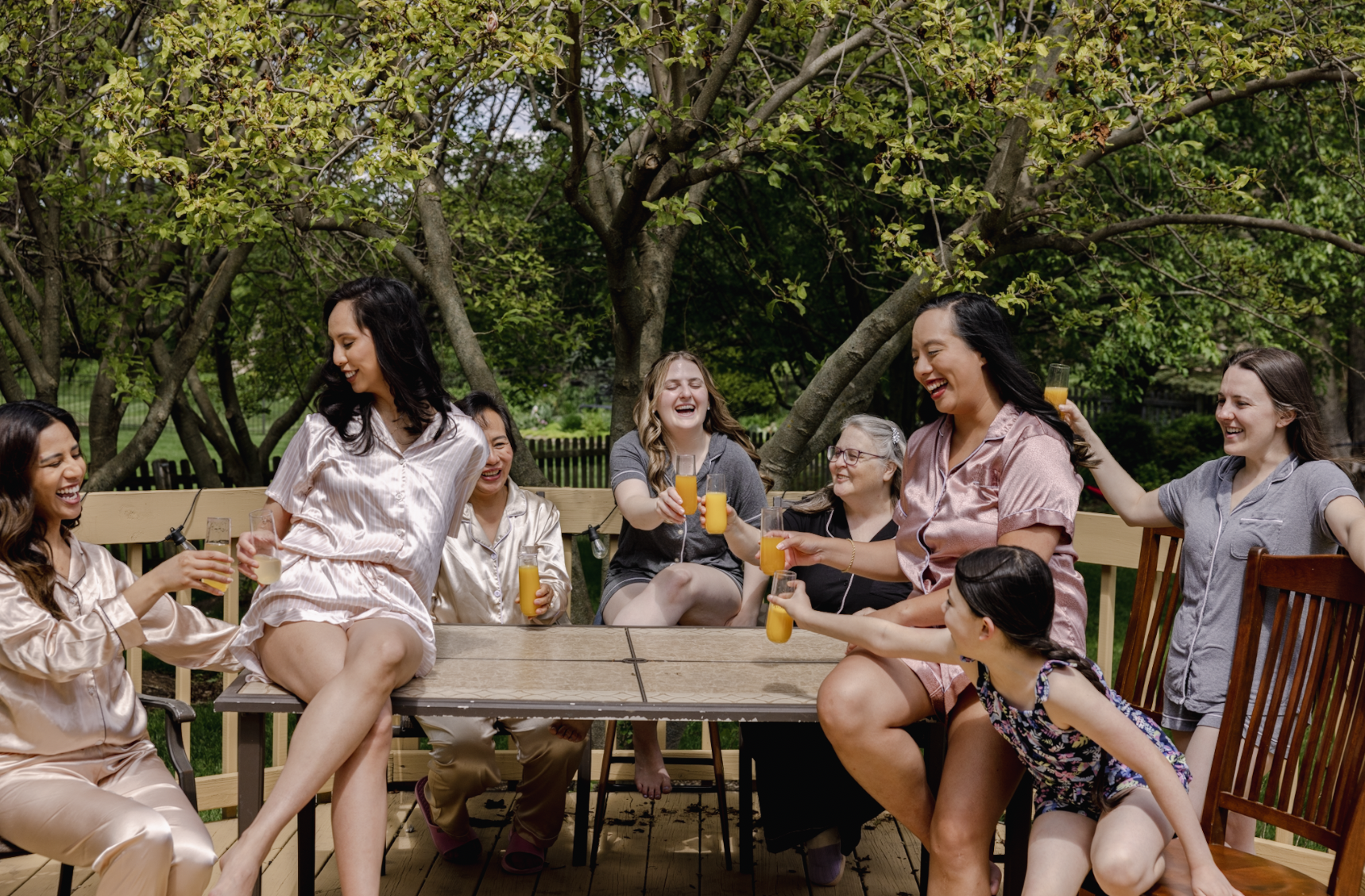 A group of women in pajamas and casual clothes enjoying drinks and laughing around a wooden table outside on a deck with trees in the background.