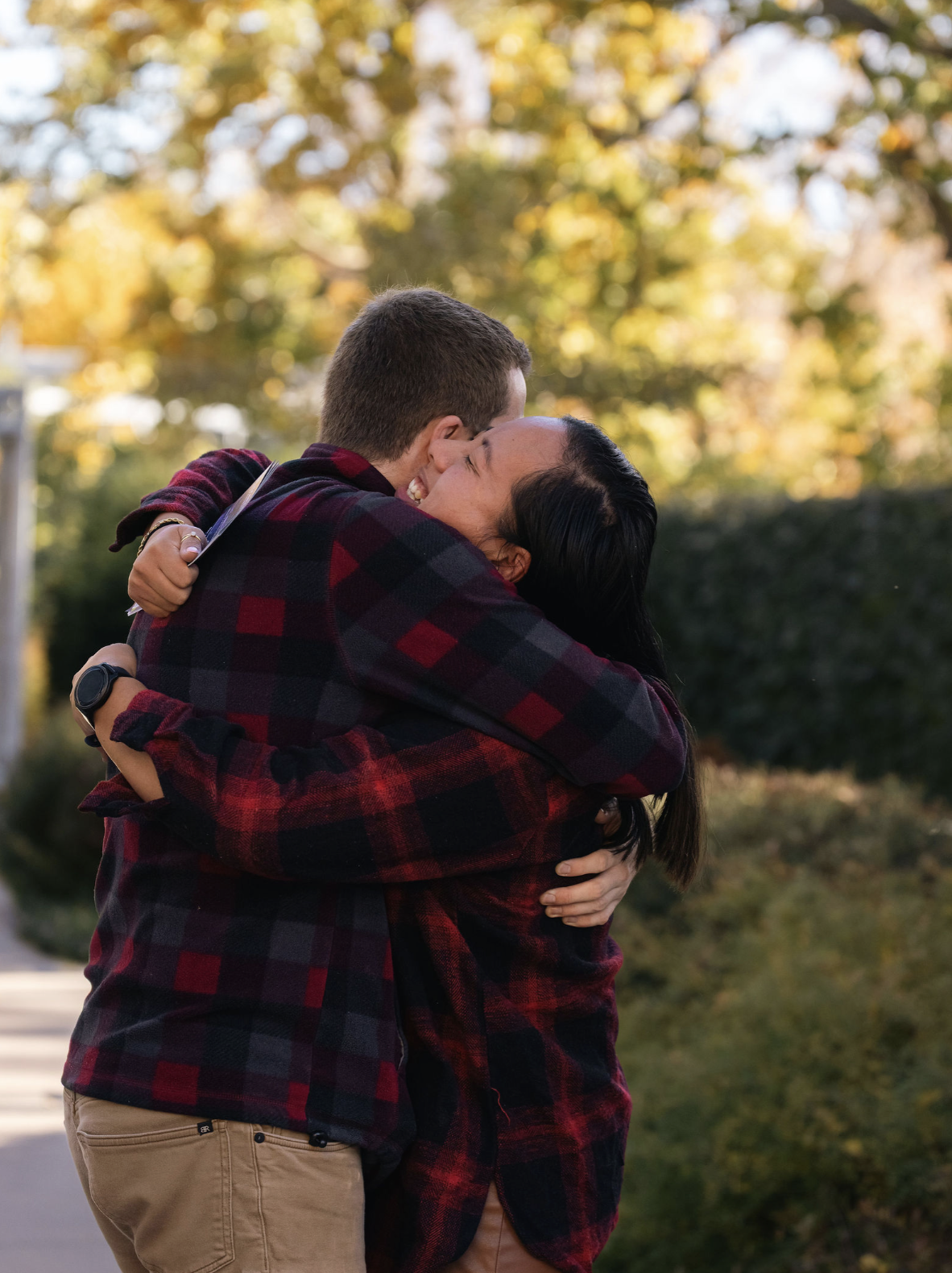 A young man and woman hugging outdoors during autumn, with trees and falling leaves in the background.