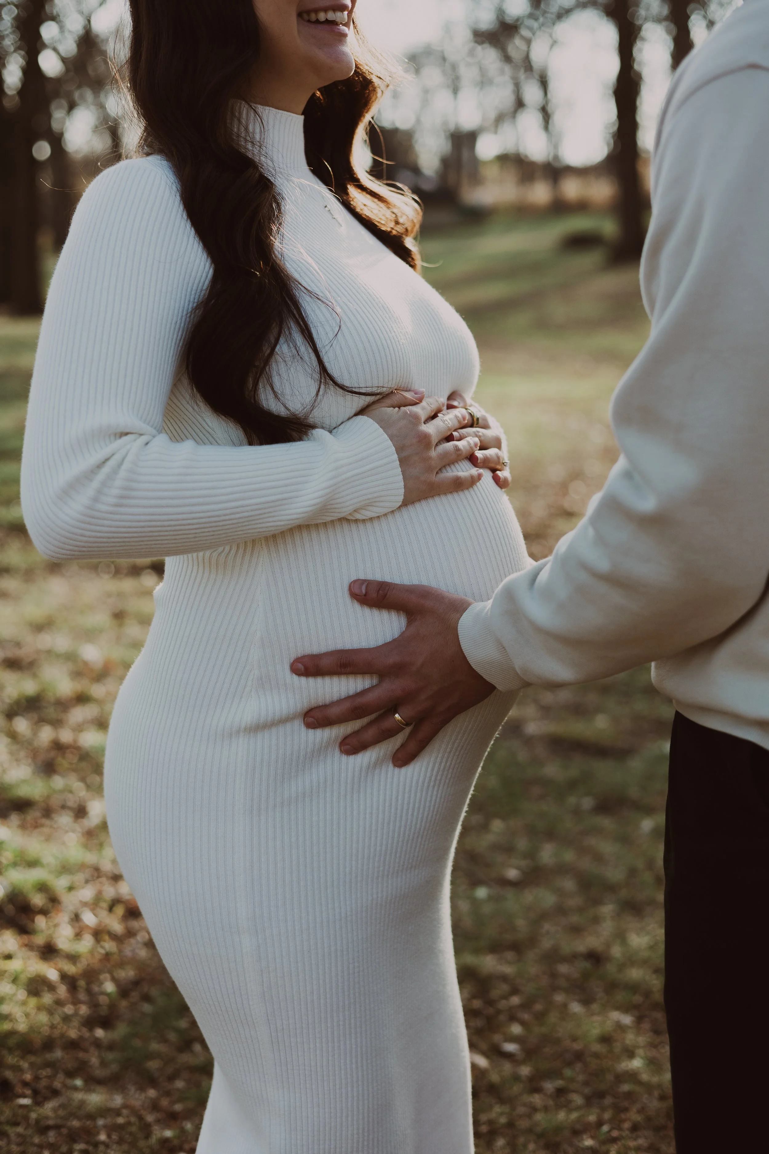 A pregnant woman in a white ribbed dress outdoors, with her hands on her belly, and a man in a white shirt gently touching her belly, both smiling and looking at each other, with trees and sunlight in the background.