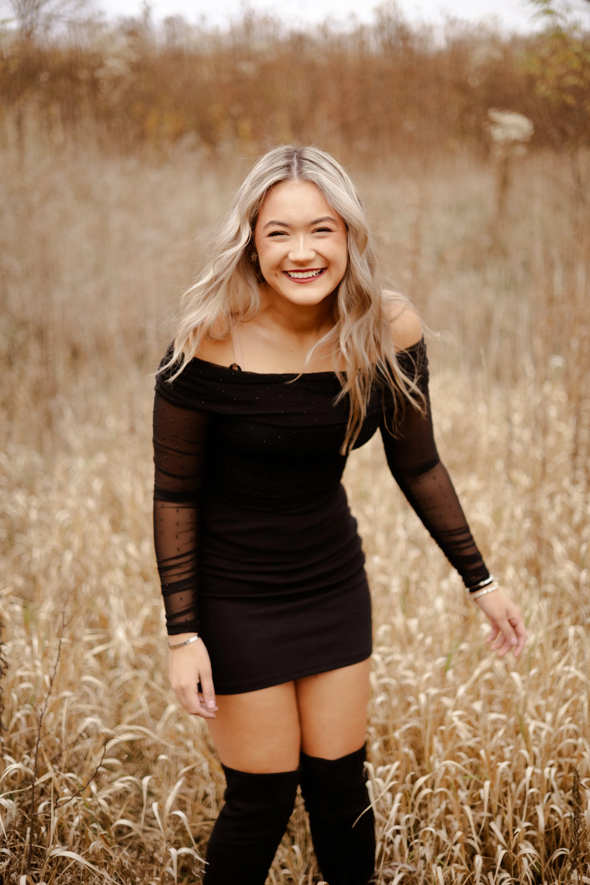 A woman with blonde hair wearing a black dress and thigh-high boots standing in a field of tall, dry grass, smiling at the camera.