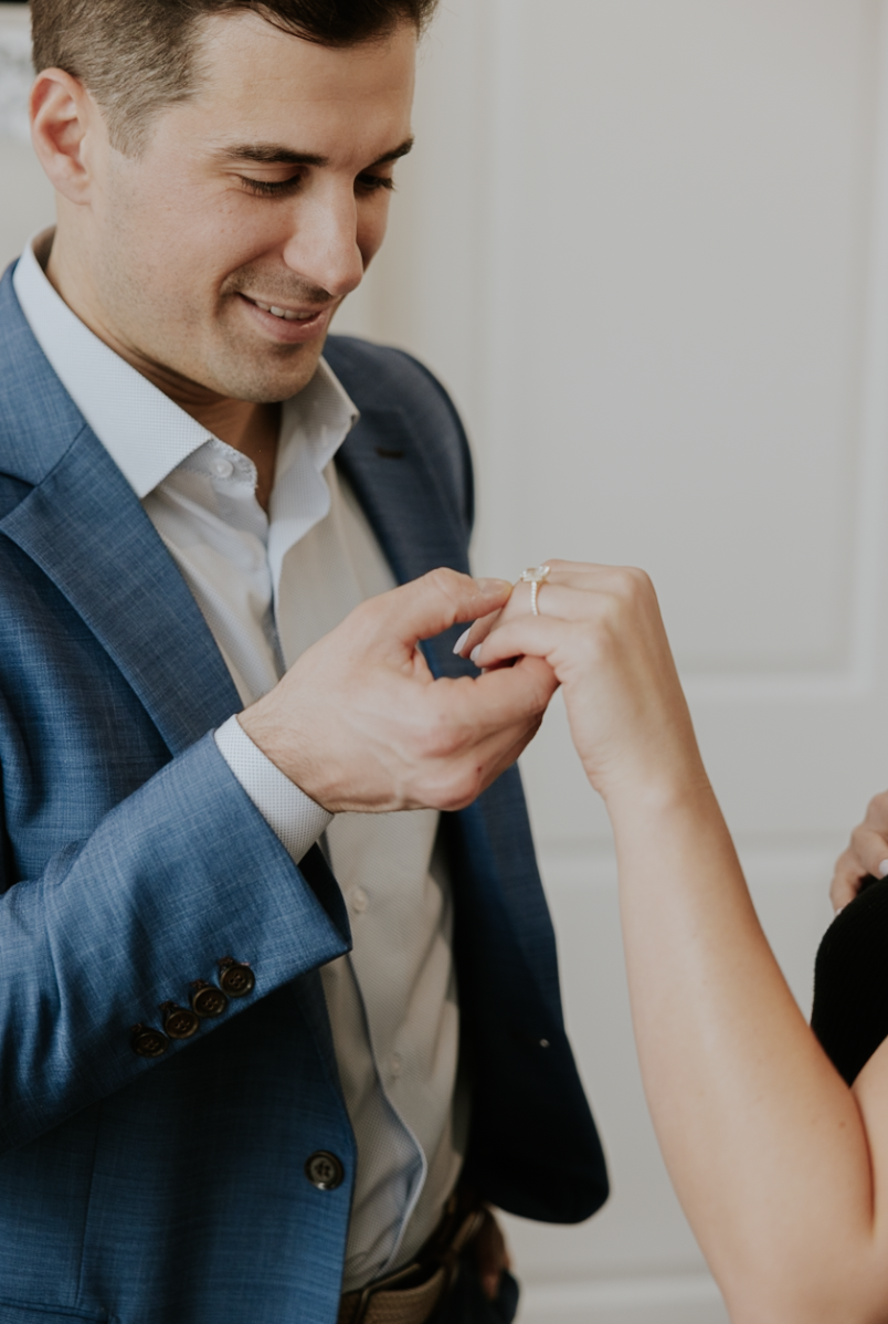 A man in a blue plaid suit looks at a woman's hand as he places a ring on her finger