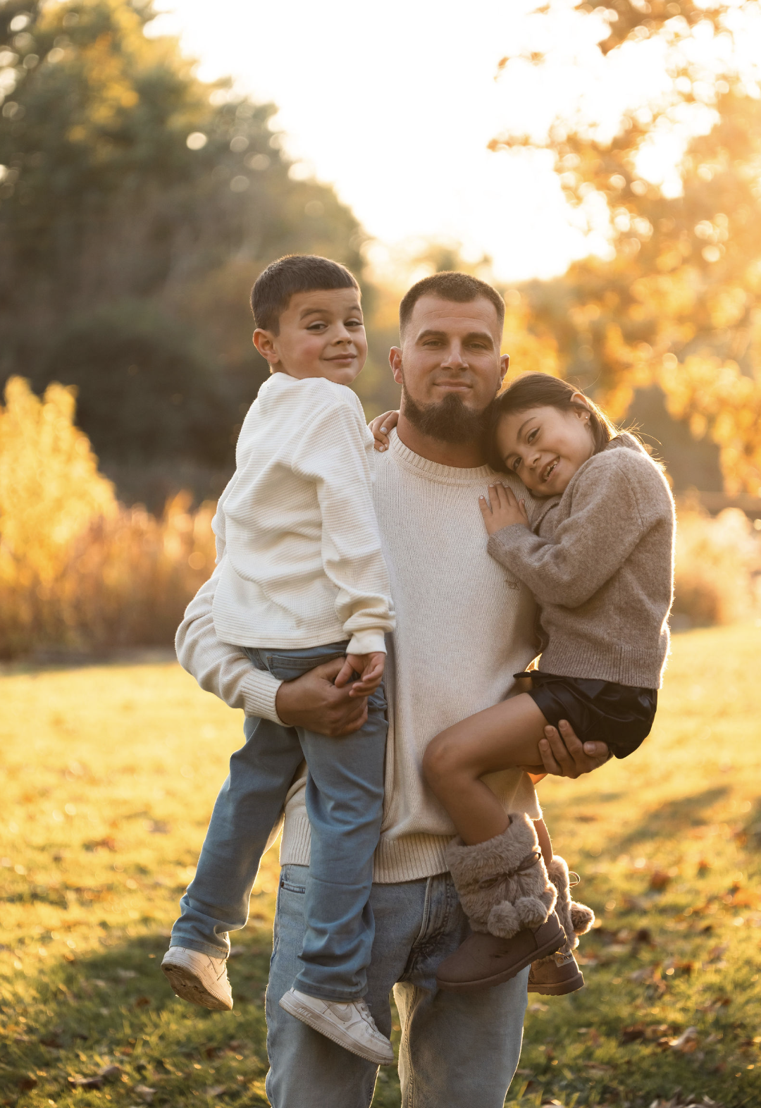 A man holding two children, a boy and a girl, outdoors during fall with golden sunlight, autumn leaves, and trees in the background.