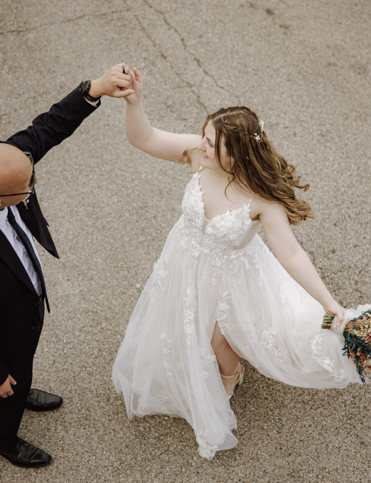 A bride with long, wavy hair and floral accessories, in a white lace wedding dress, dancing with a man in a black suit and glasses, on a gravel surface, holding hands.
