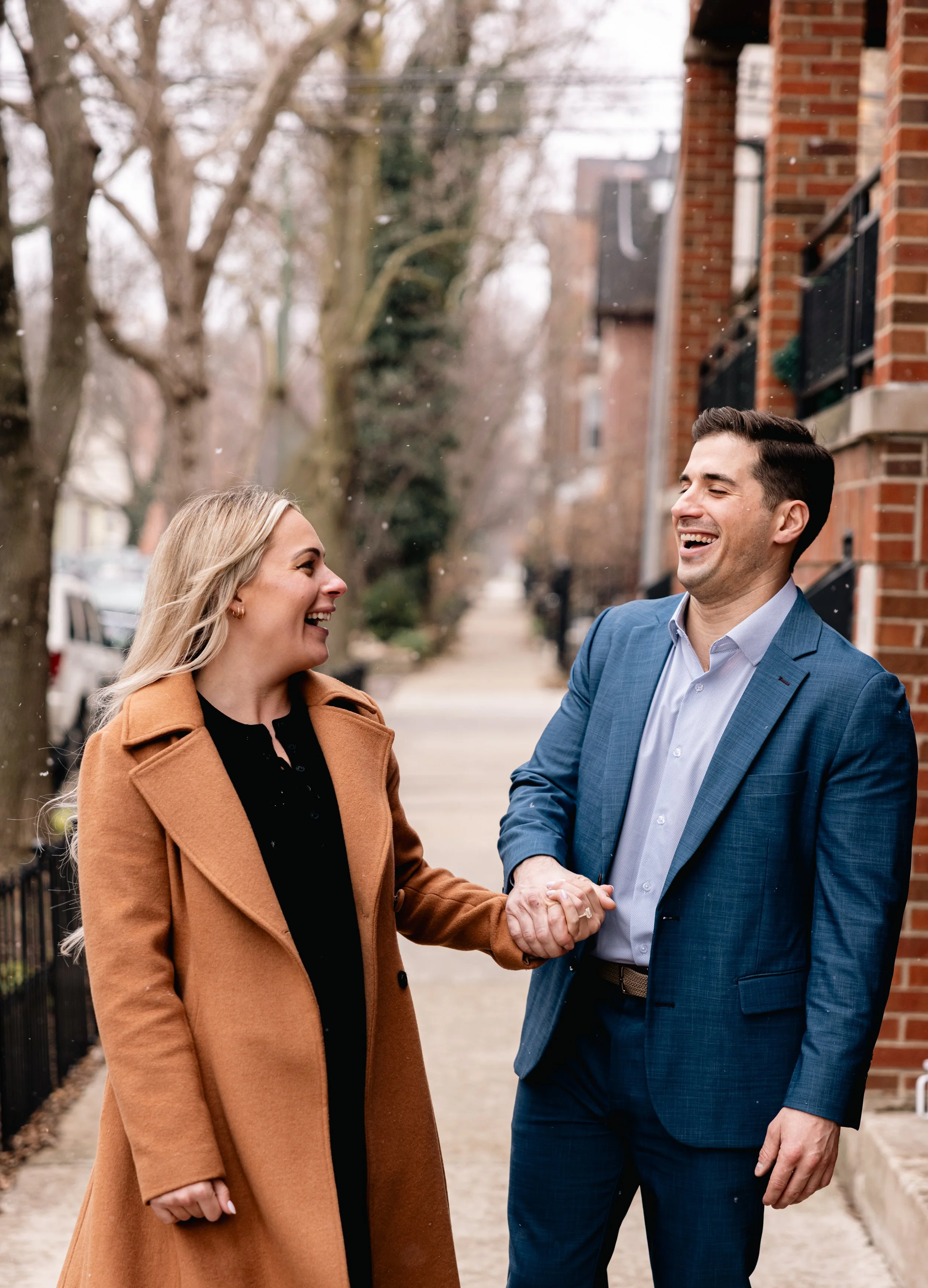 A man and a woman shaking hands outdoors on a sidewalk, smiling and looking at each other, with leafless trees and brick buildings in the background.