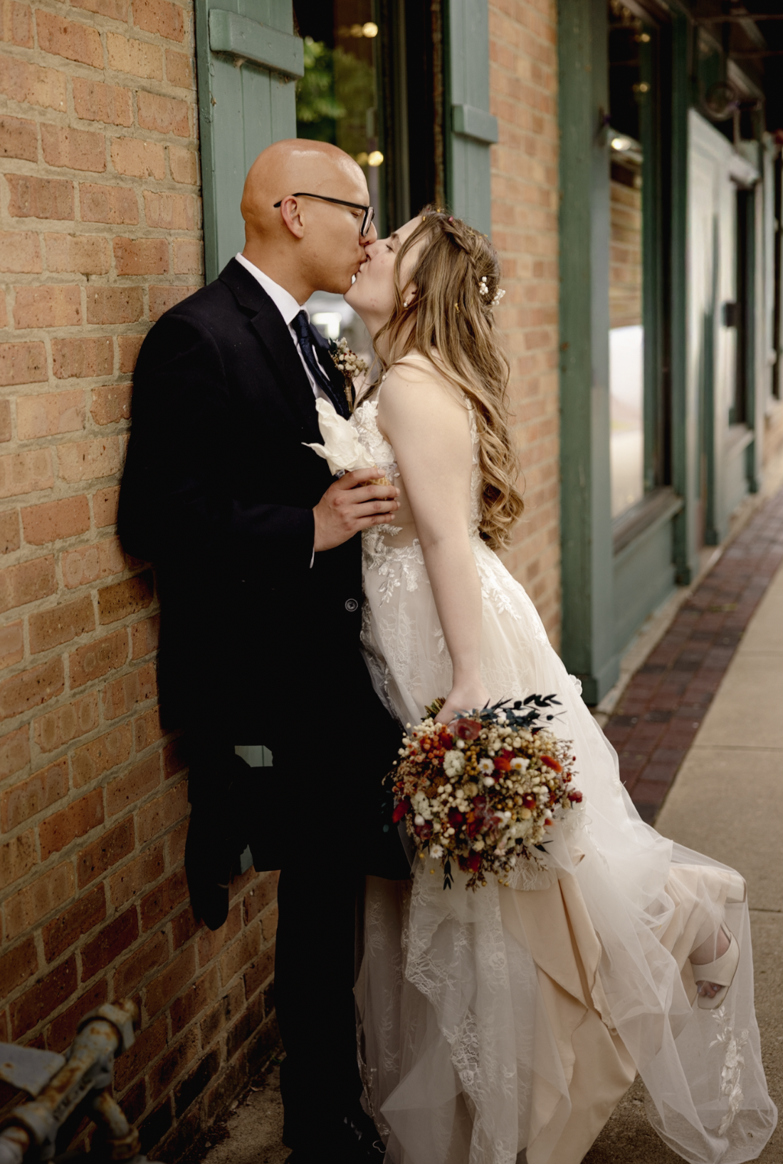 A newlywed couple kissing against a brick wall on a city sidewalk, the bride holding a colorful bouquet of flowers.