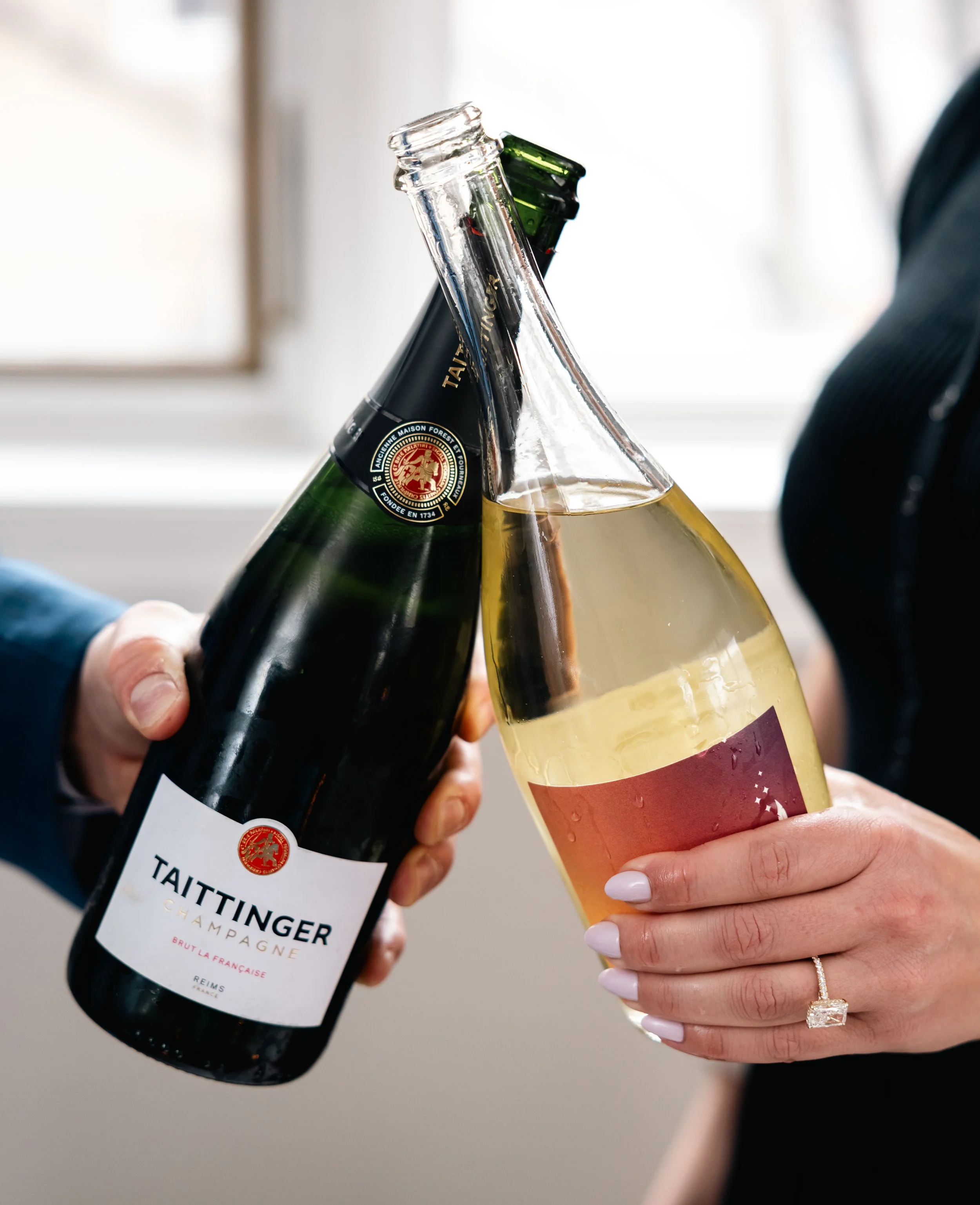 Two people clinking wine bottles and glasses in a toast, with a blurry background and casual attire.