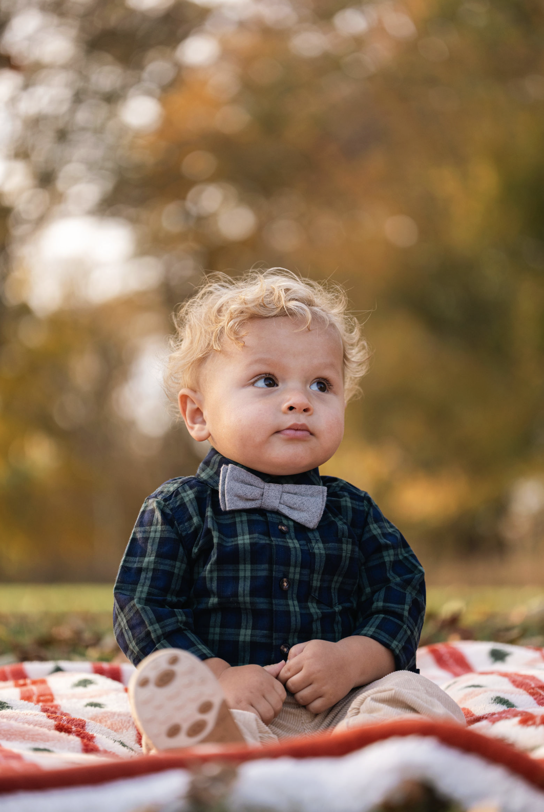 A young child with curly blonde hair sitting on a red and white checkered blanket outdoors during autumn, wearing a plaid shirt, gray bowtie, beige pants, and brown shoes, with fall foliage blurred in the background.