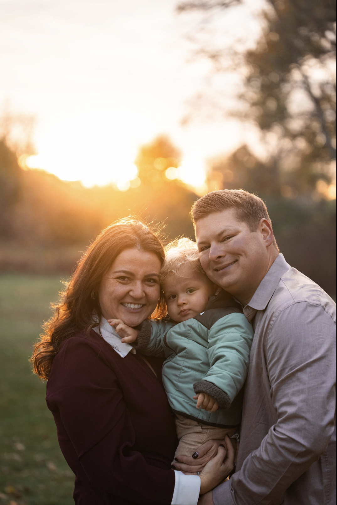 Family of three outdoors during sunset, smiling, with trees in the background.