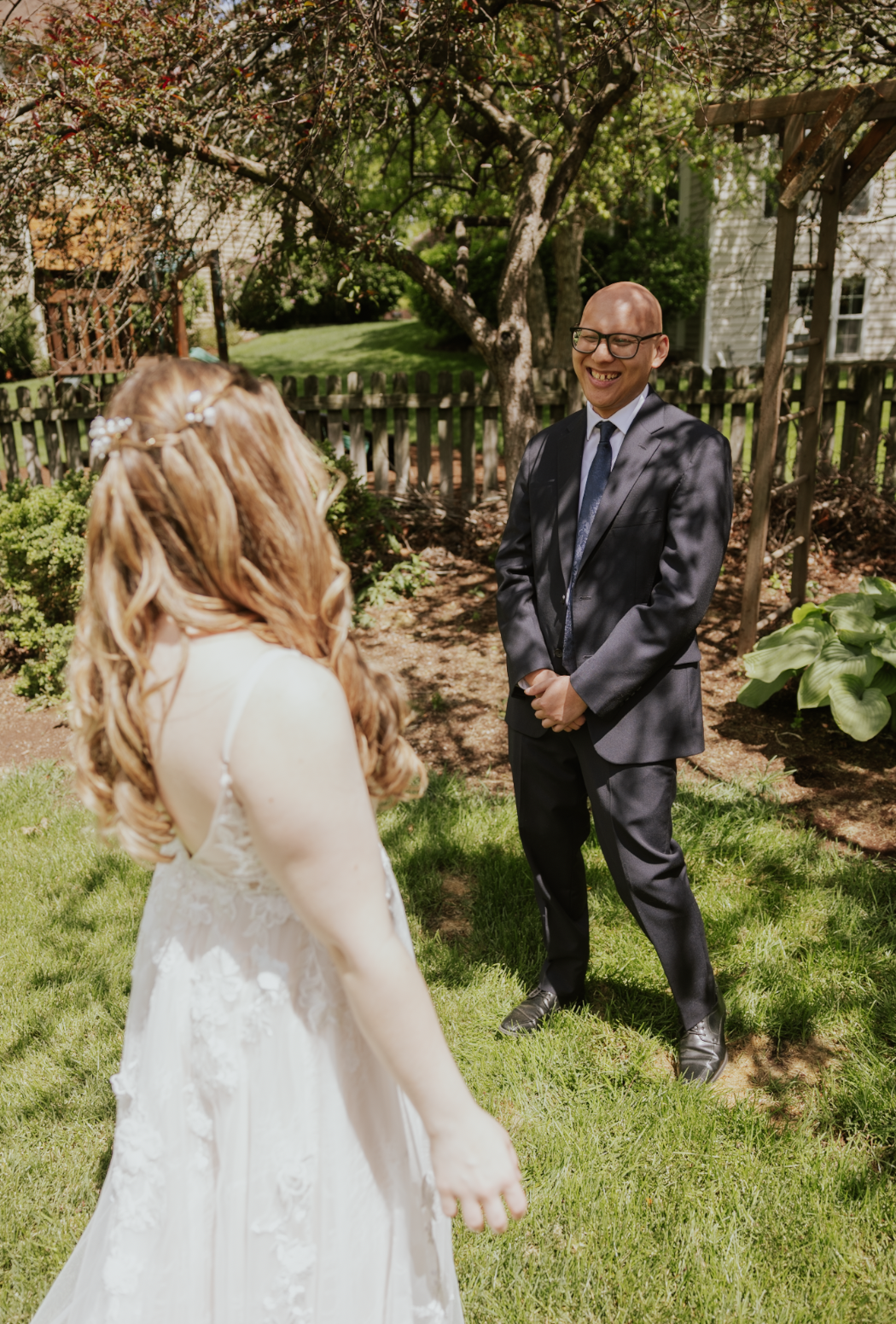 A man in a suit smiling and standing in a garden with a woman in a wedding dress, who has curly red hair, during daytime.