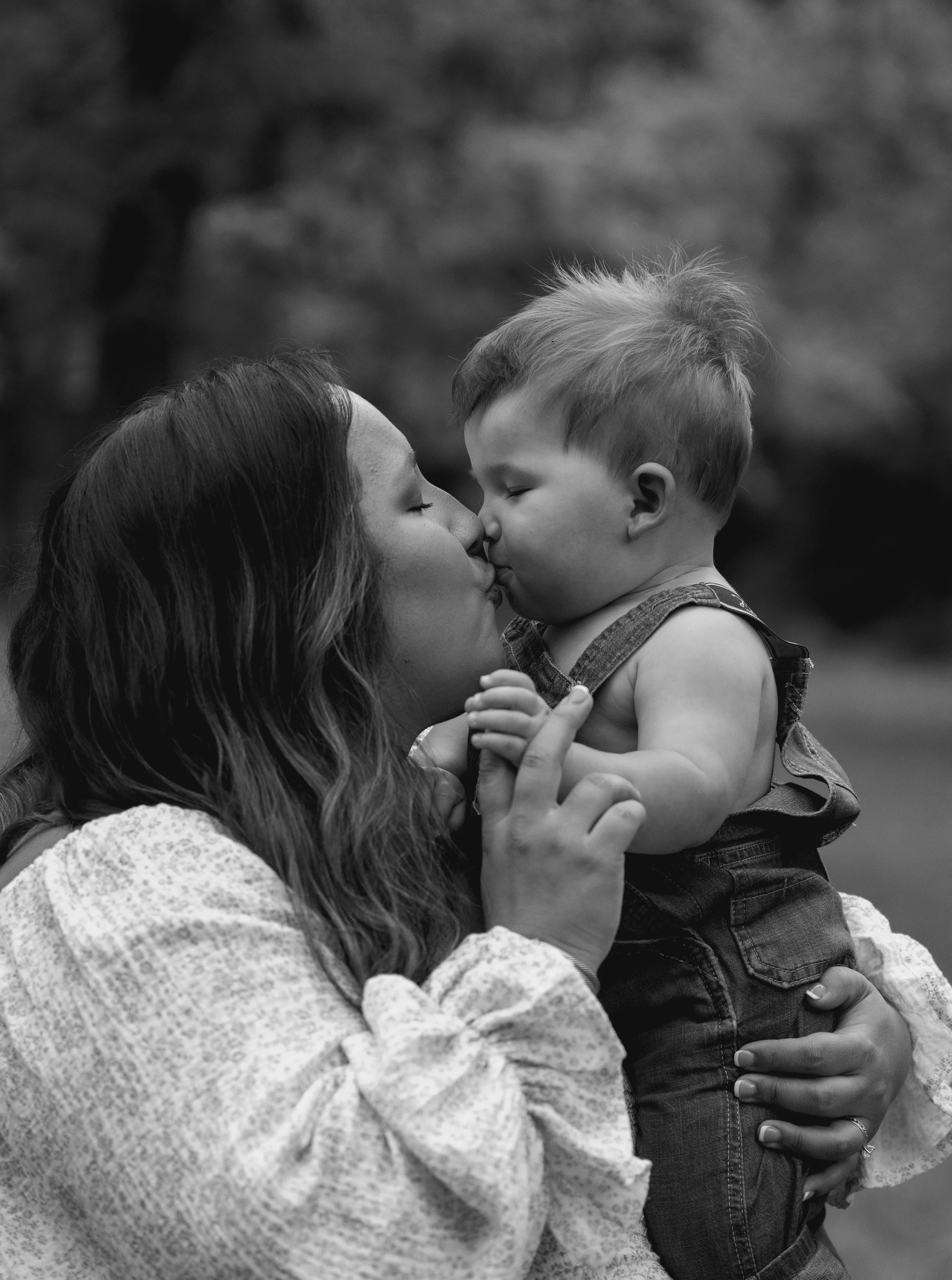 A woman with long hair and a light-colored sweater is kissing a young boy on the nose. The boy, wearing a sleeveless shirt and jeans, is being held by the woman, with his eyes closed, in an outdoor setting with blurred trees in the background.