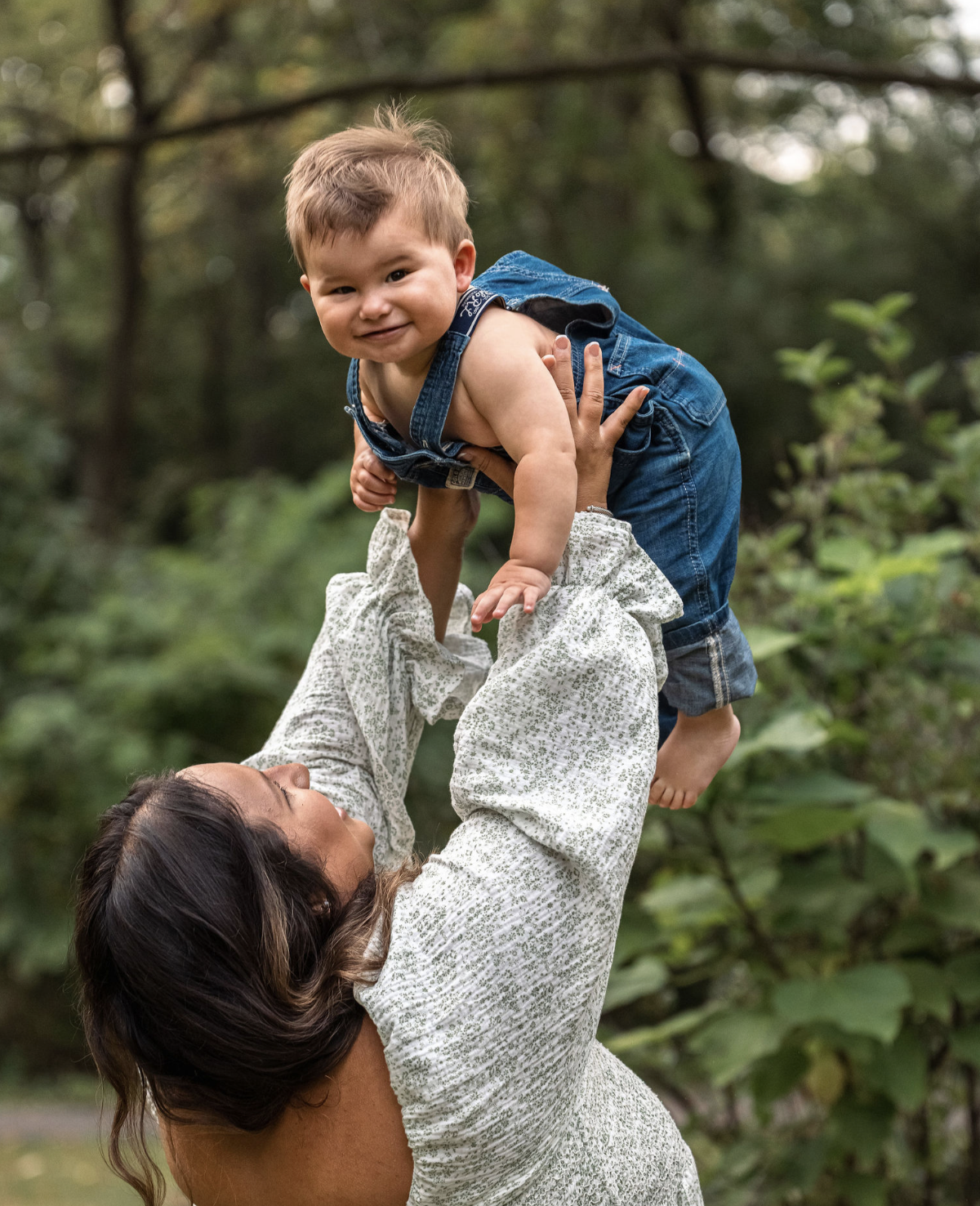 A woman is lifting a young boy in the air outdoors in a green, wooded area. The boy is smiling and looking at the camera, wearing denim overalls, while the woman has dark hair and is wearing a light-colored, patterned shirt.