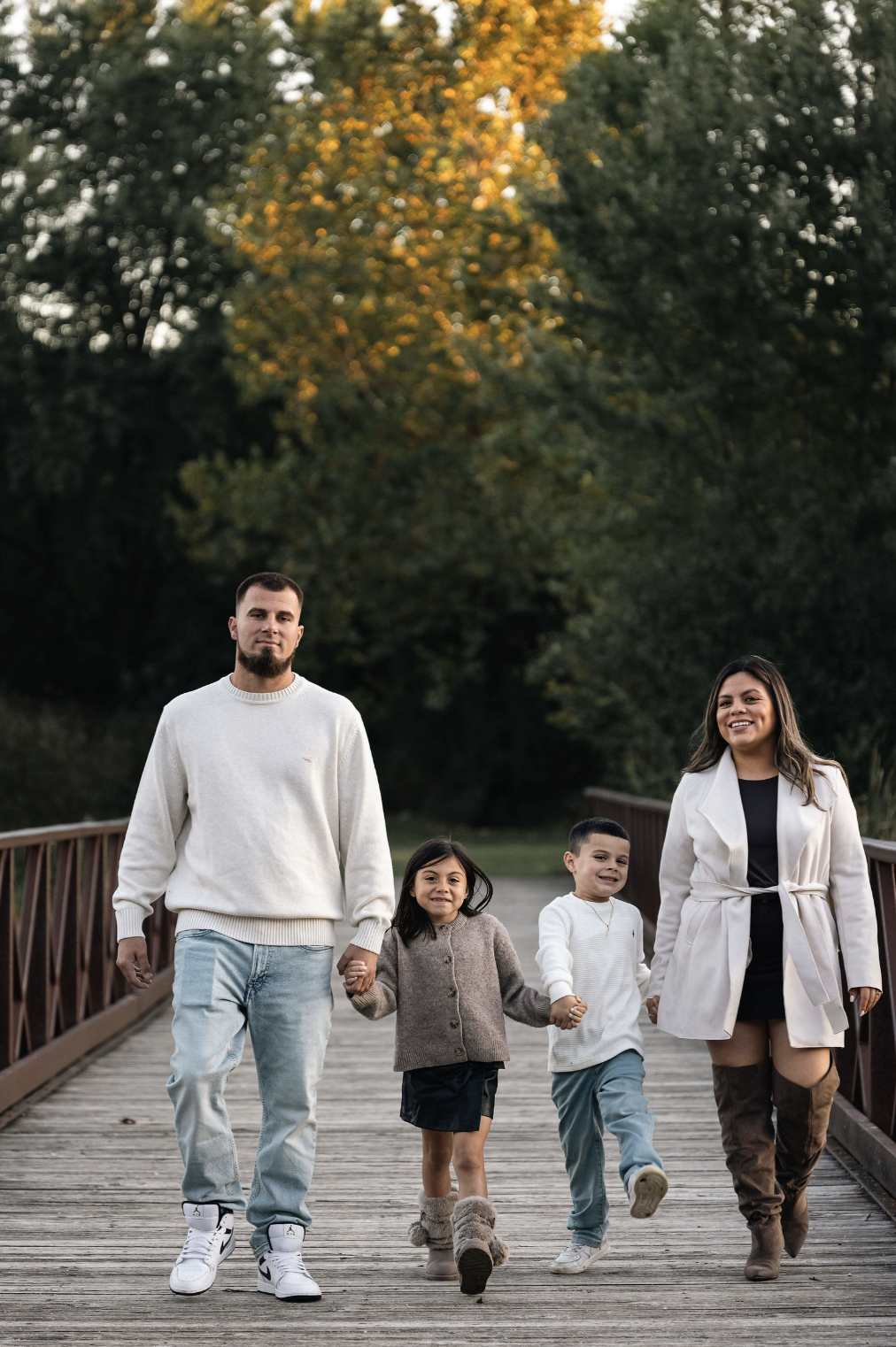A family of four walking on a wooden bridge outdoors during autumn, holding hands, with trees showing yellow and green foliage in the background.