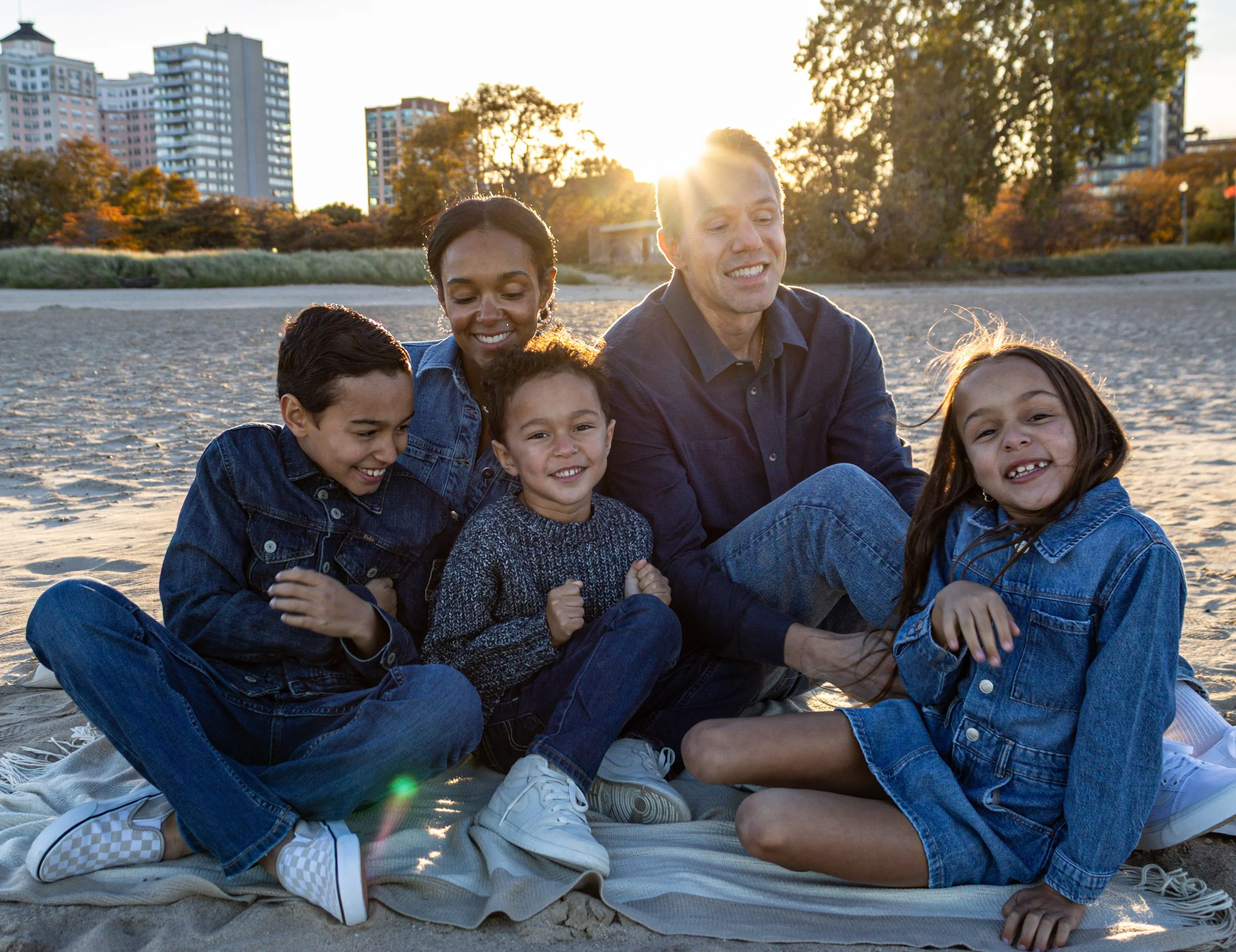 A happy multiracial family of six sitting on a blanket at the beach during sunset, with high-rise buildings and trees in the background.