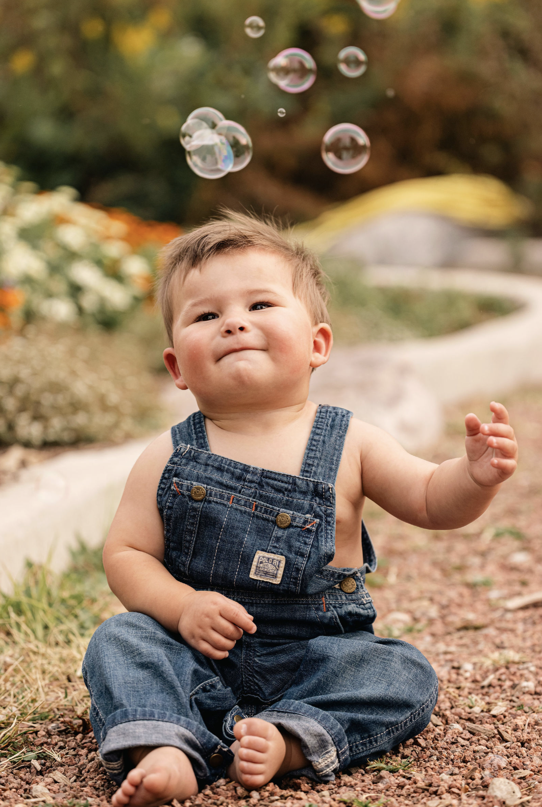 A young kid sitting outdoors on a gravel path, playing with soap bubbles. The child is wearing denim overalls and no shirt, with a content expression. In the background, there are colorful flowering plants and blurred greenery.