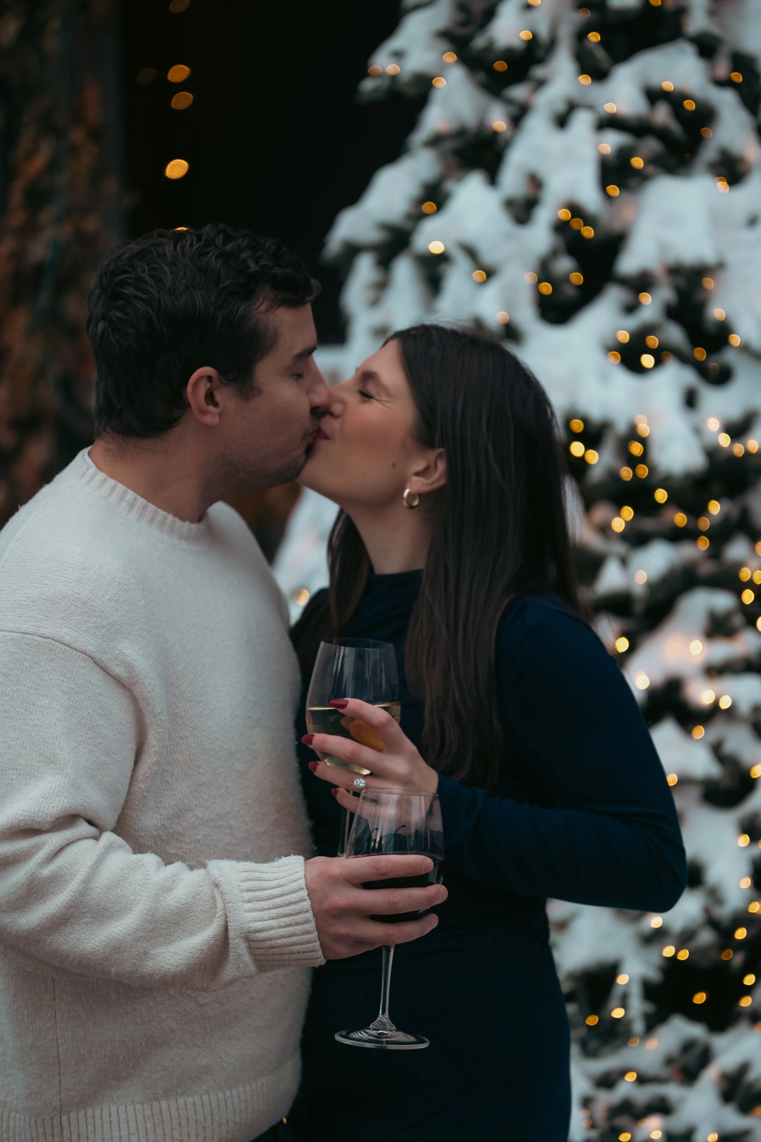 A man and woman sharing a kiss during a festive celebration near a decorated Christmas tree, with the woman holding a glass of white wine.