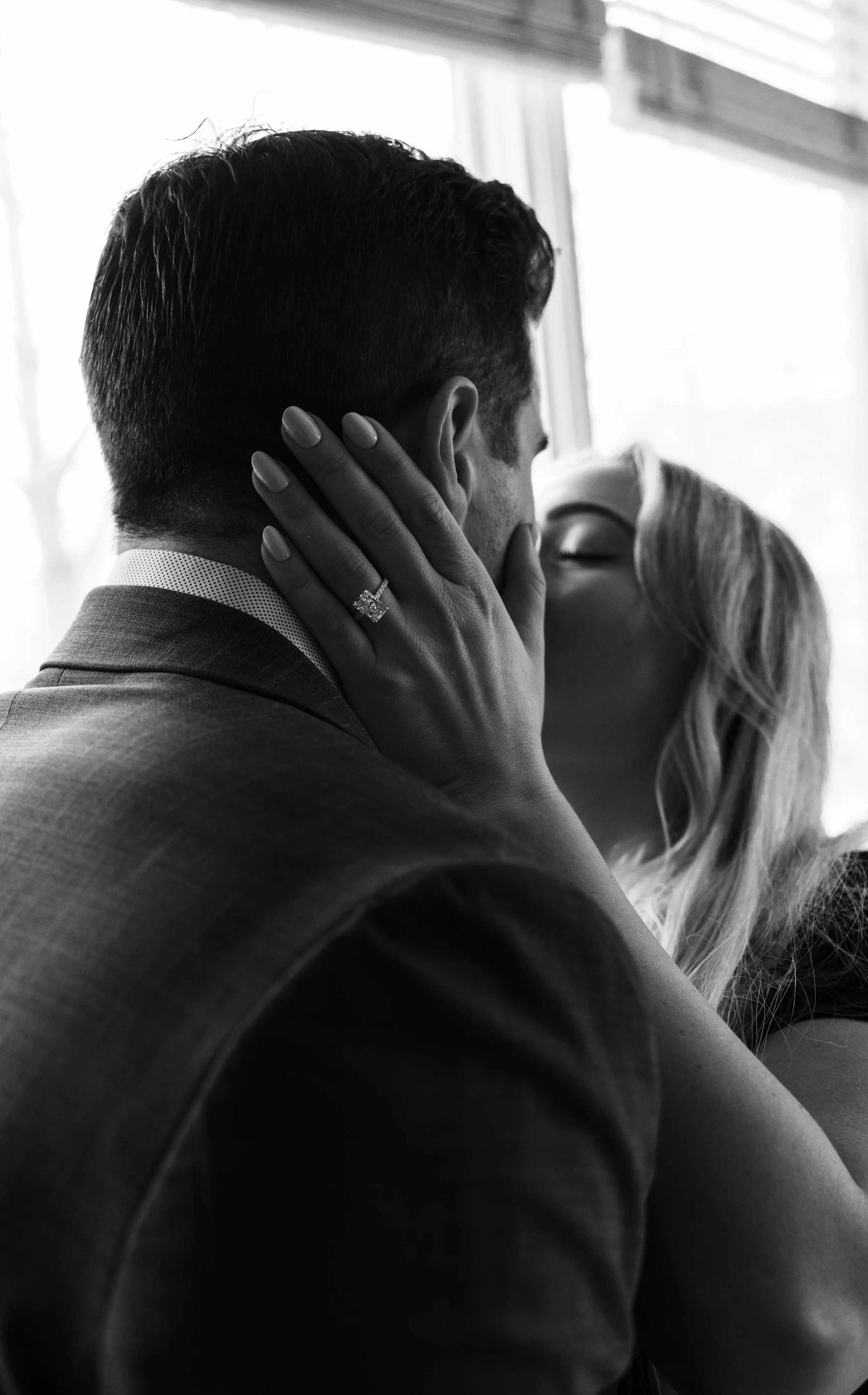 A black and white photograph of a couple sharing an intimate moment, with the woman gently holding the man's face and their faces close together, near a window.