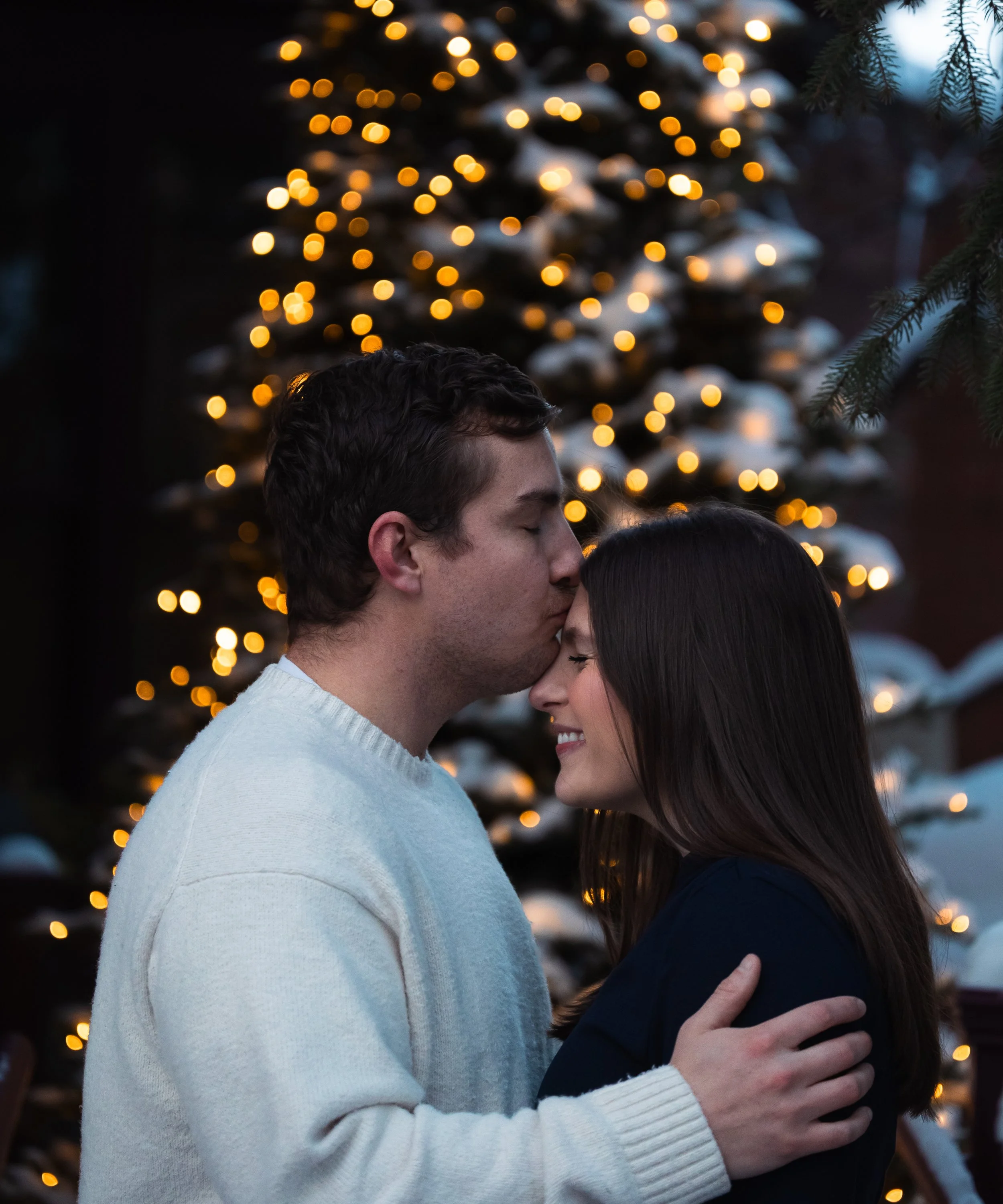 A couple sharing a kiss in front of a Christmas tree with blurred lights in the background.