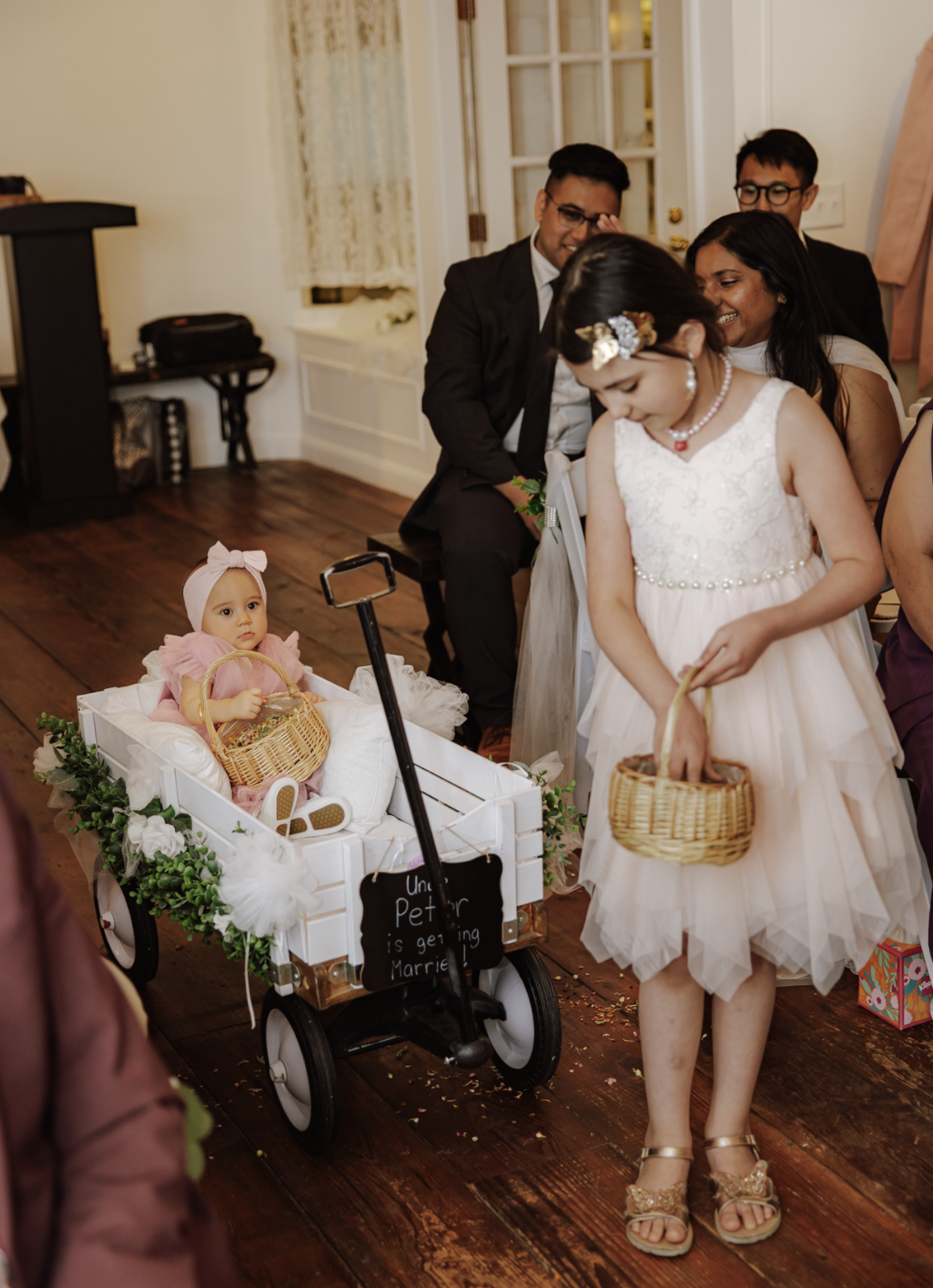 A young girl in a white dress with pearl accessories and a floral headband is holding a small basket, walking near a doll in a white cart with a sign that says ''Uncle Peter is getting married.'' Other people are seated nearby, and some are smiling a