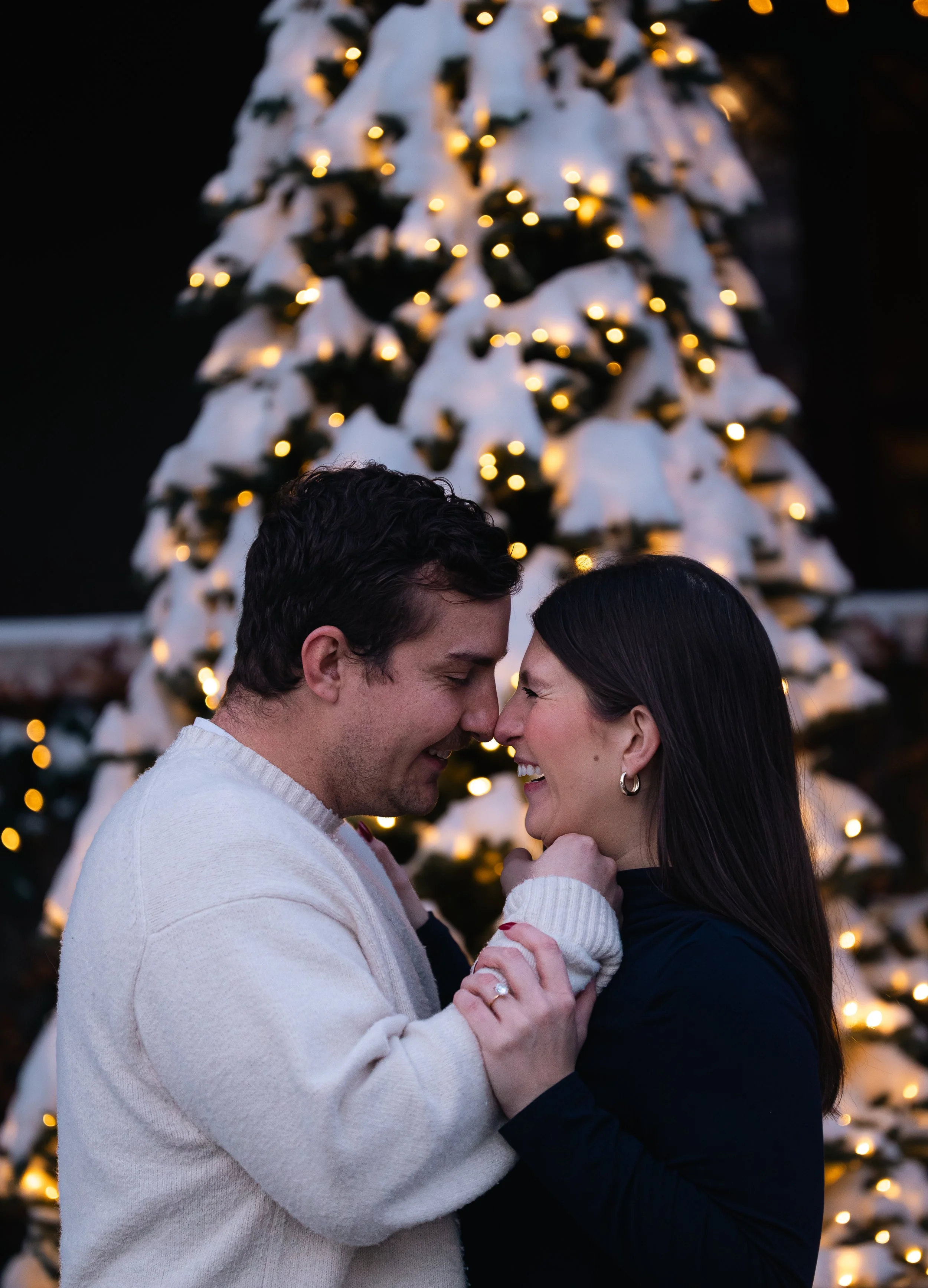 A couple faces each other closely, touching foreheads and smiling, during a Christmas celebration with a decorated Christmas tree with lights in the background.