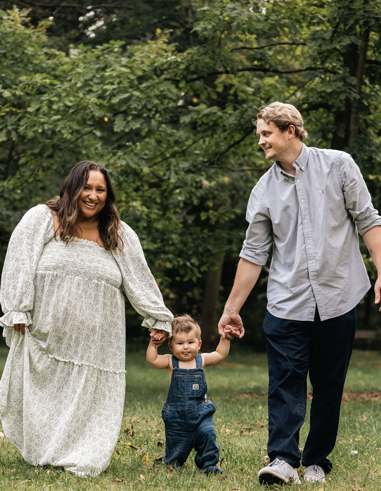 A family of three with a woman, a man, and a young child walking outdoors in a park with green trees in the background. The woman is wearing a white dress, the man is in a gray shirt and jeans, and the child is in overalls.