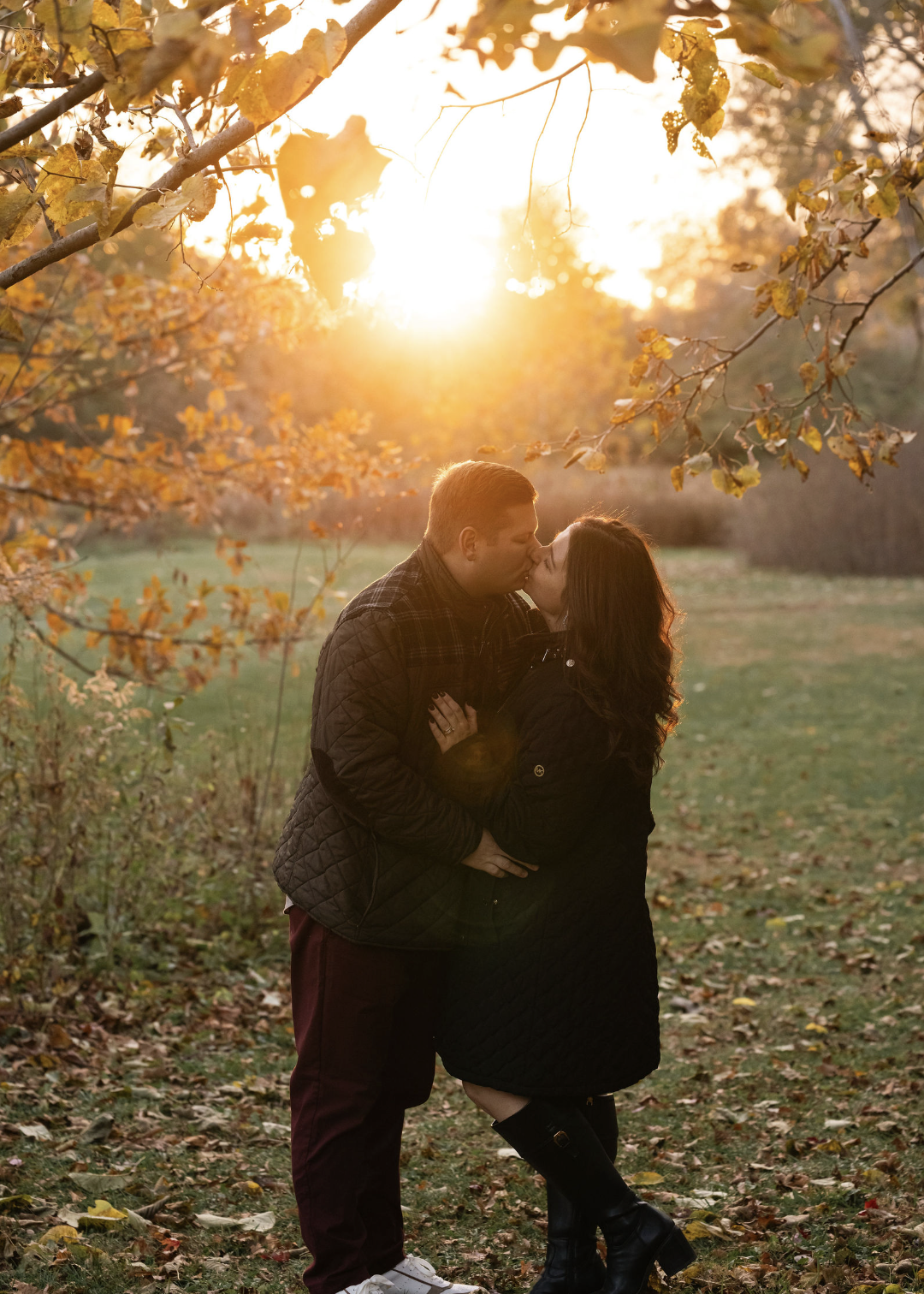 A couple sharing a kiss outdoors in an autumn setting with trees and fallen leaves, during sunset.