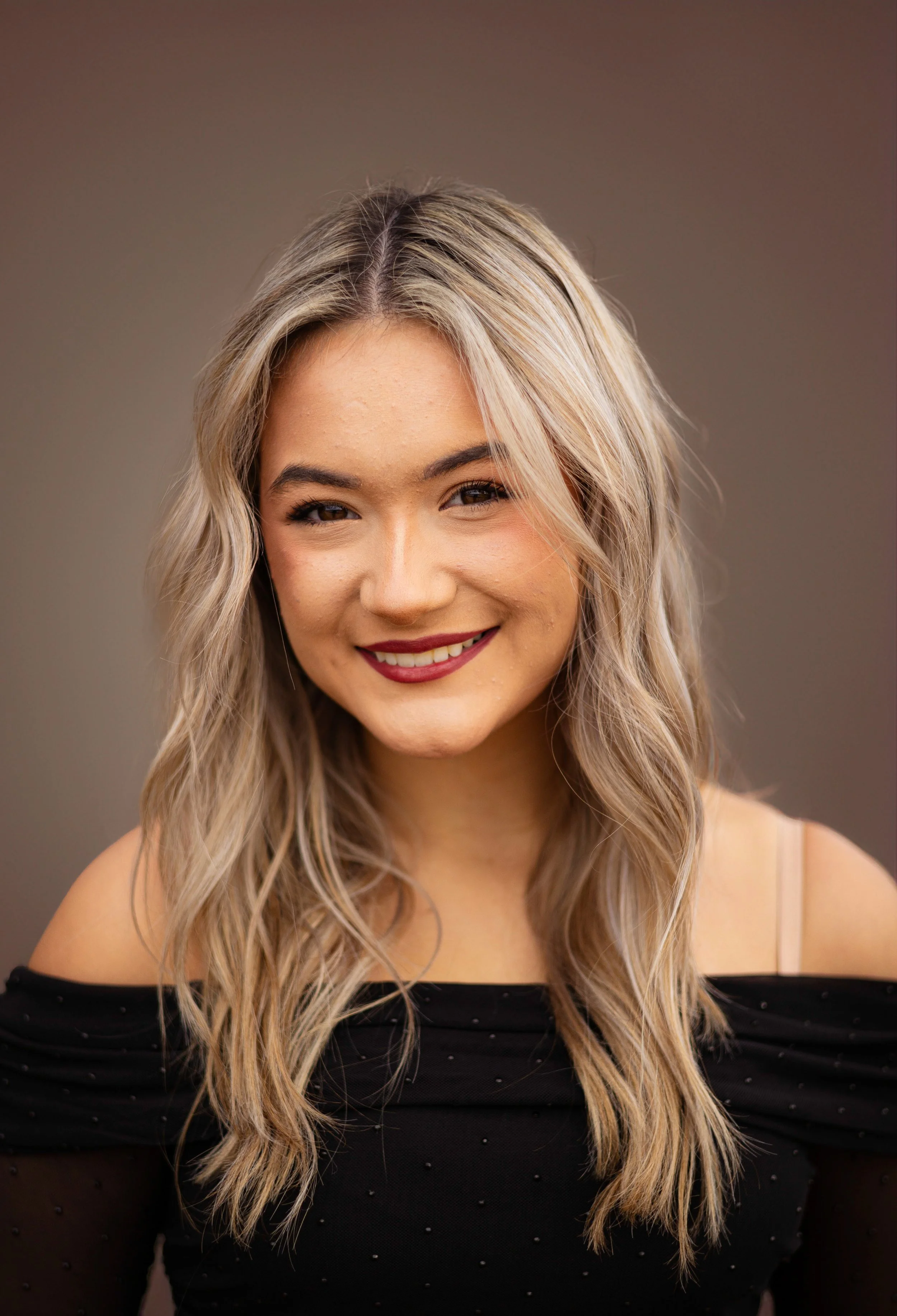 A young woman with blonde wavy hair smiling at the camera. She wears a black off-the-shoulder top with small embellishments.
