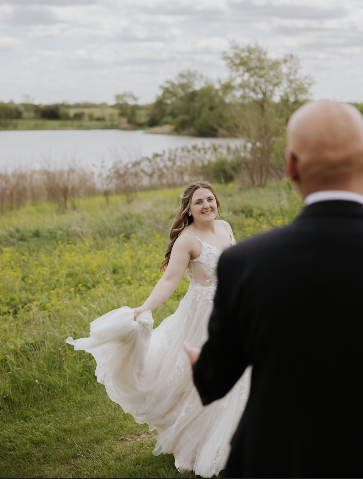 A bride in a white wedding gown smiling and holding her dress in a grassy field, with a man in a black suit in the foreground, near a lake and trees under a cloudy sky.