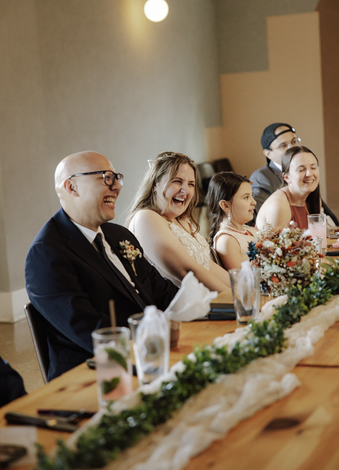 People sitting at a long decorated table, laughing and smiling, during a wedding reception, with floral centerpieces and drinks in front of them.