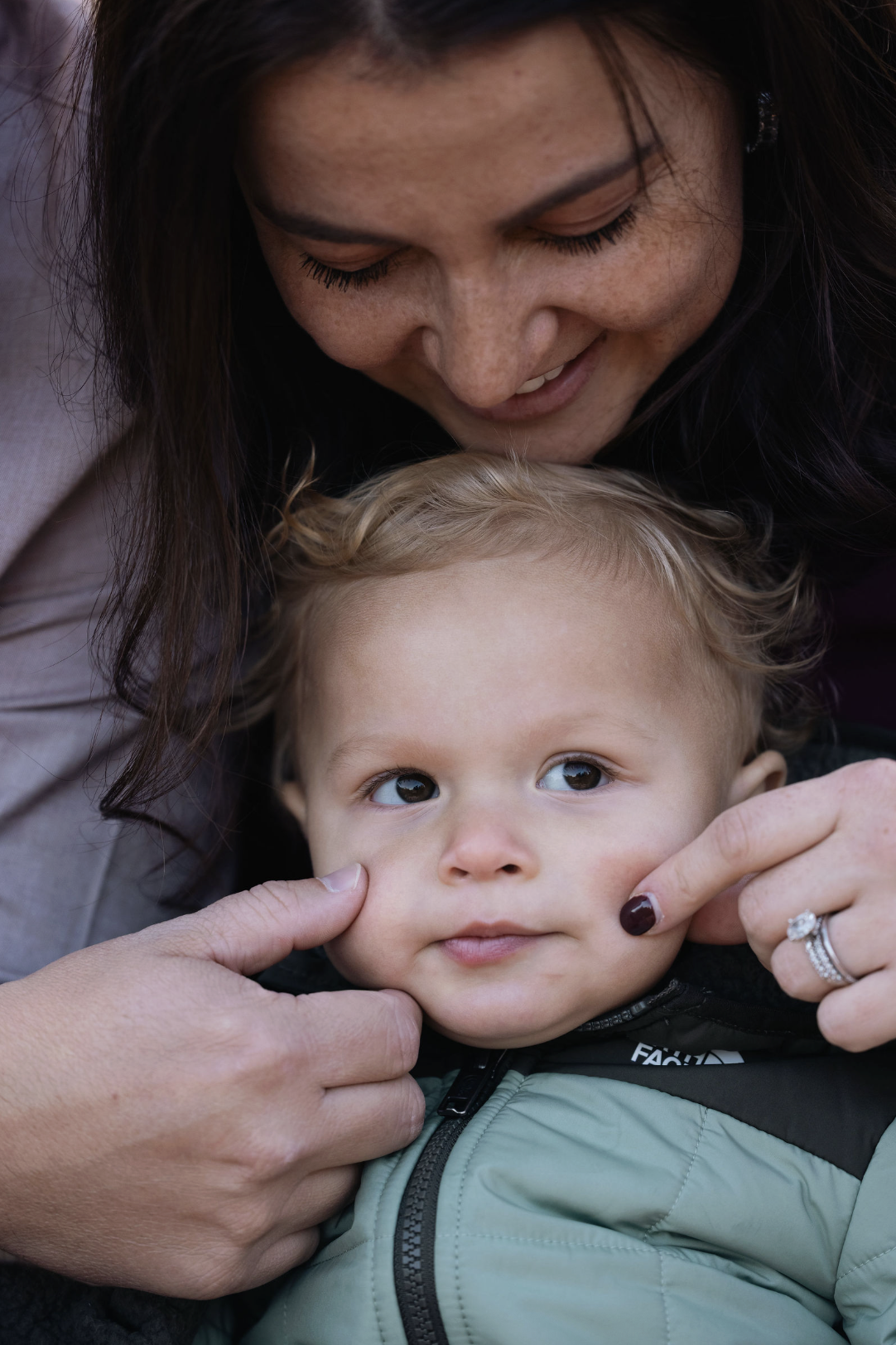 A woman with long dark hair smiling and leaning over a young child with blond hair, who is looking up and being gently pinched on the cheeks by her hands.