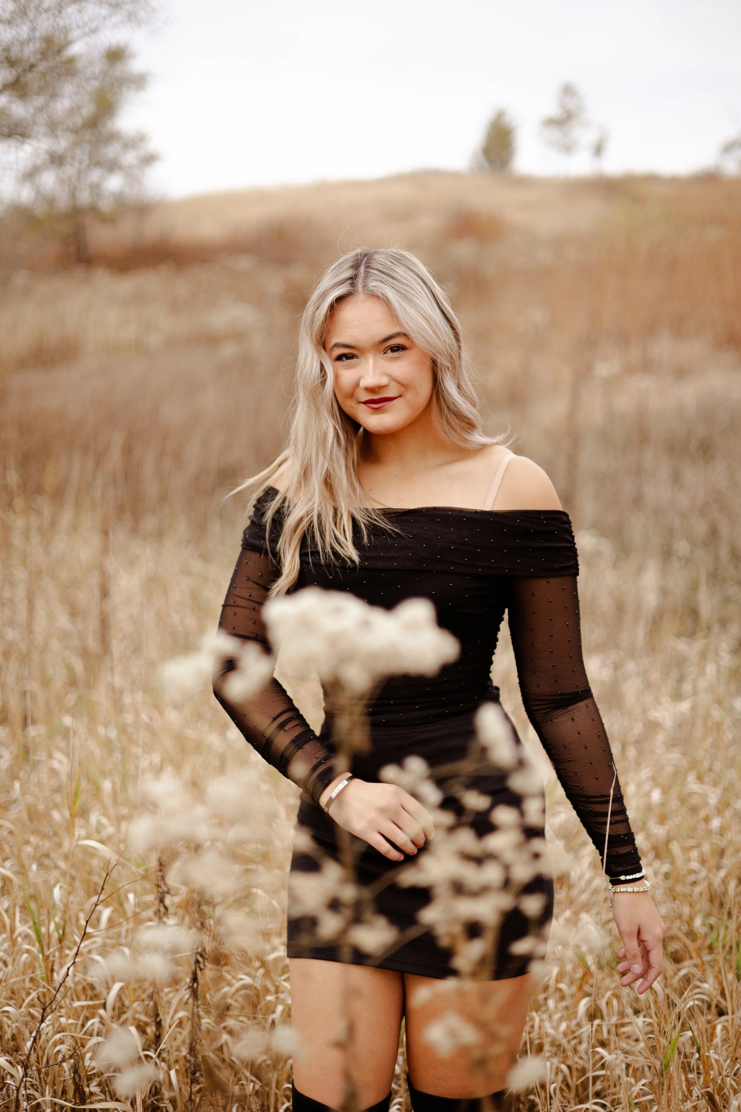 A woman with long blonde hair in a black off-shoulder dress standing in a field of tall dry grass with a blurred hillside in the background.
