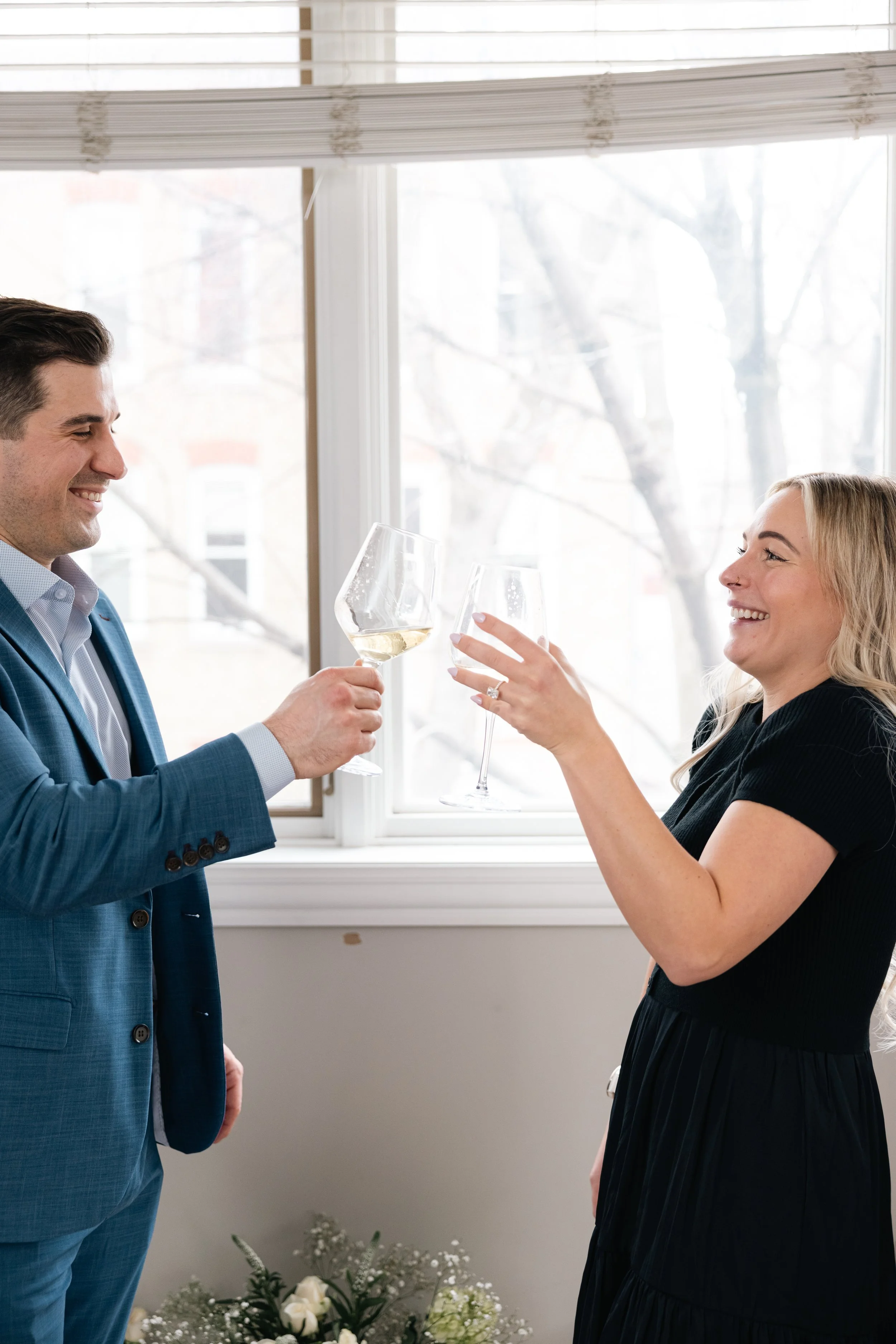 A man and woman sharing a toast with glasses of white wine indoors near a window with blinds, with flowers at the bottom of the window.