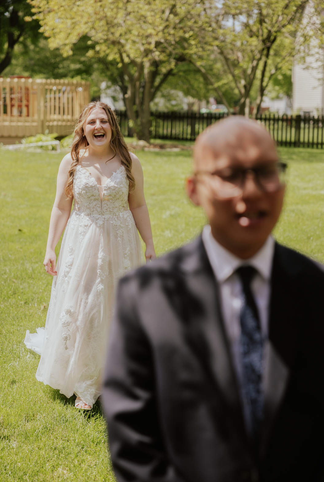 A woman in a white lace wedding dress is smiling and walking towards a man in a suit, outdoors in a backyard with green grass, trees, and a wooden fence.
