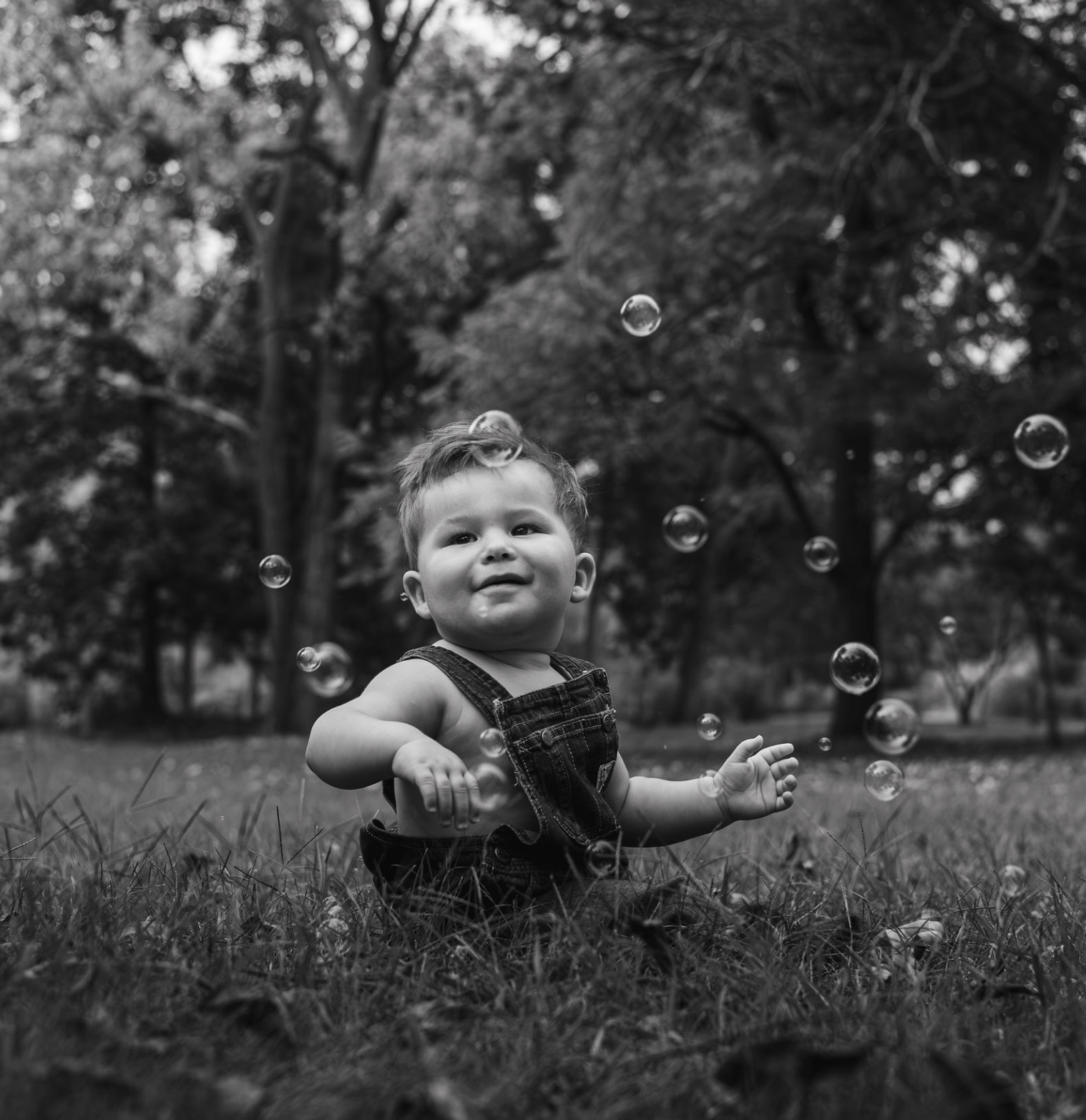 Black and white photo of a young child sitting on grass outdoors, surrounded by floating bubbles with trees in the background.