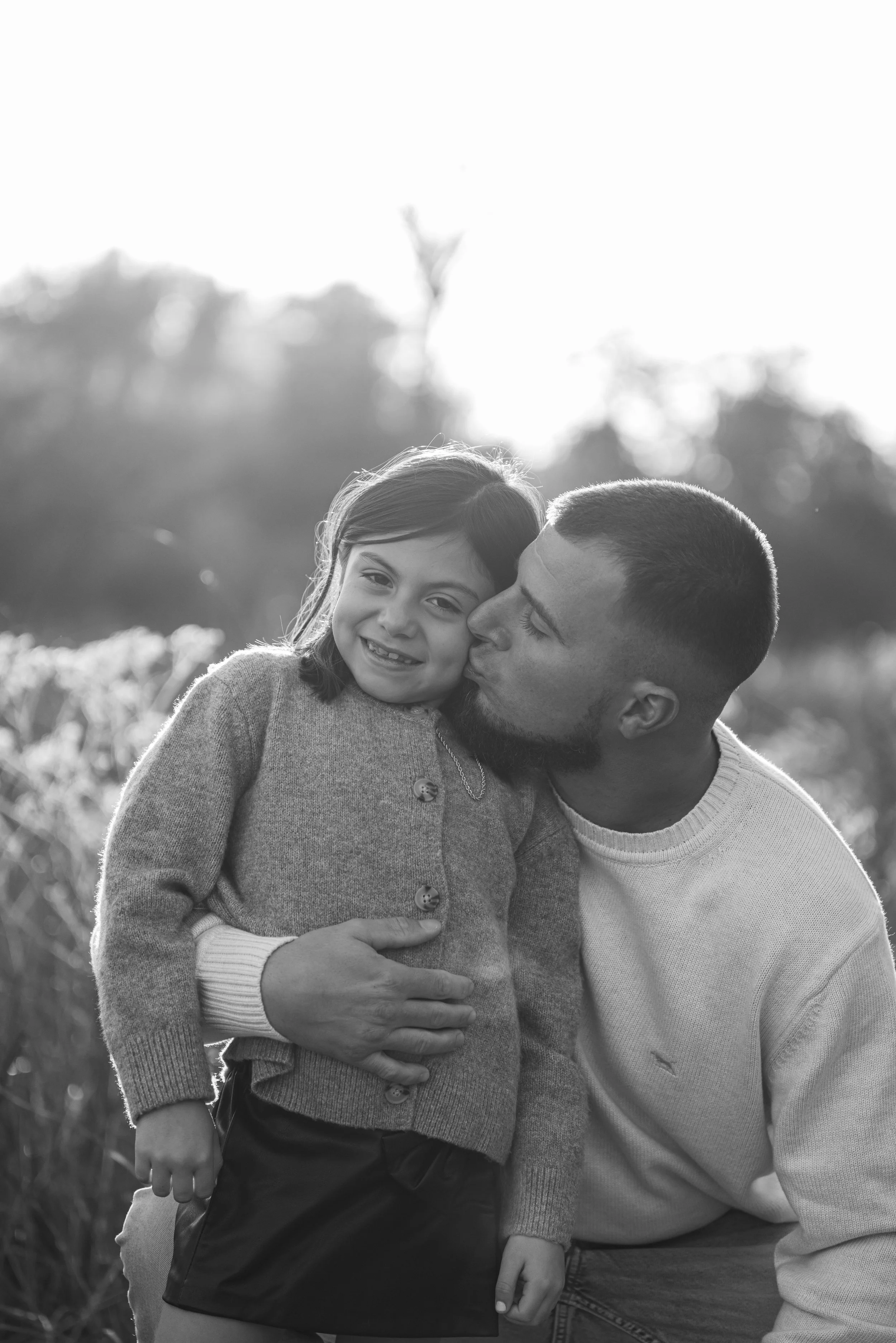 A man and a girl sharing a kiss and hug outdoors, with a blurred natural background in black and white.