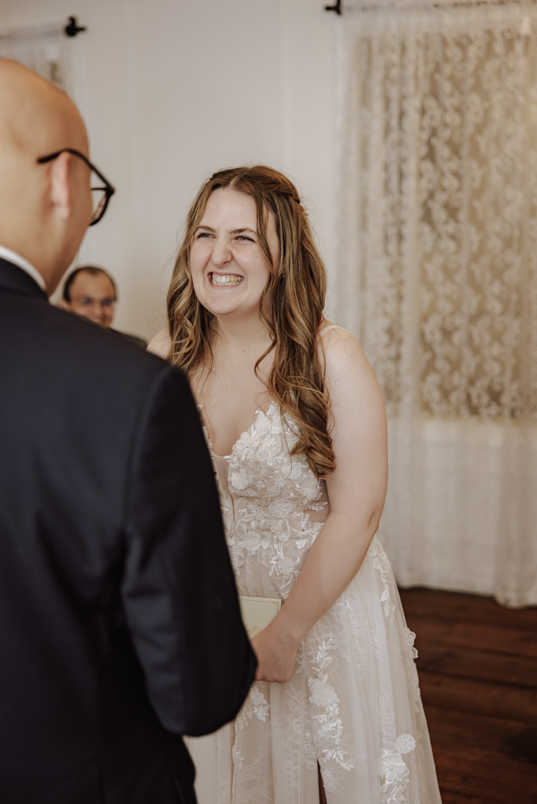 A bride with long, wavy brown hair wearing a white lace wedding dress, smiling with her teeth showing, during her wedding ceremony. An officiant or groom is partially visible on the left.