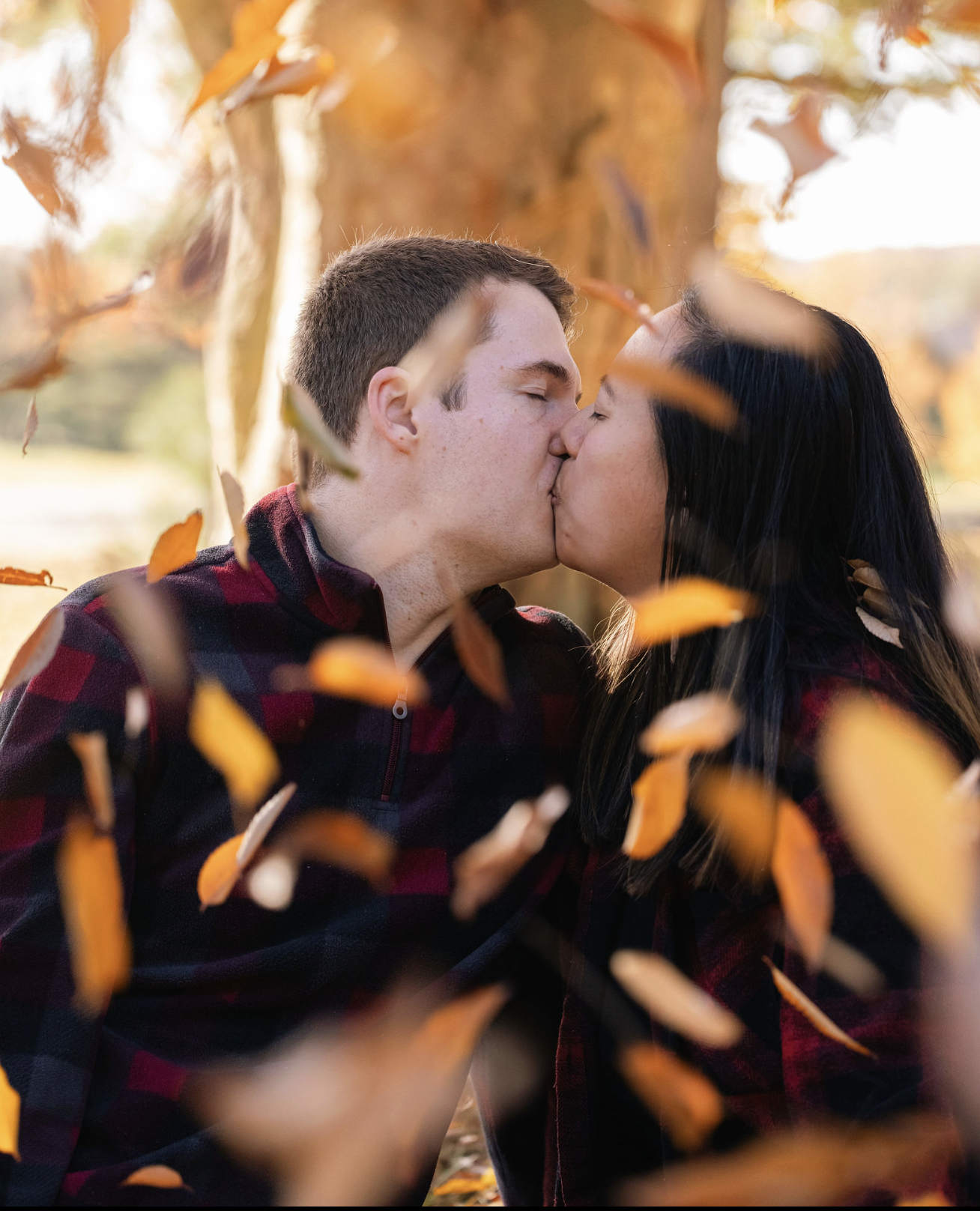 A young couple sharing a kiss outdoors, surrounded by falling autumn leaves, with trees in the background.