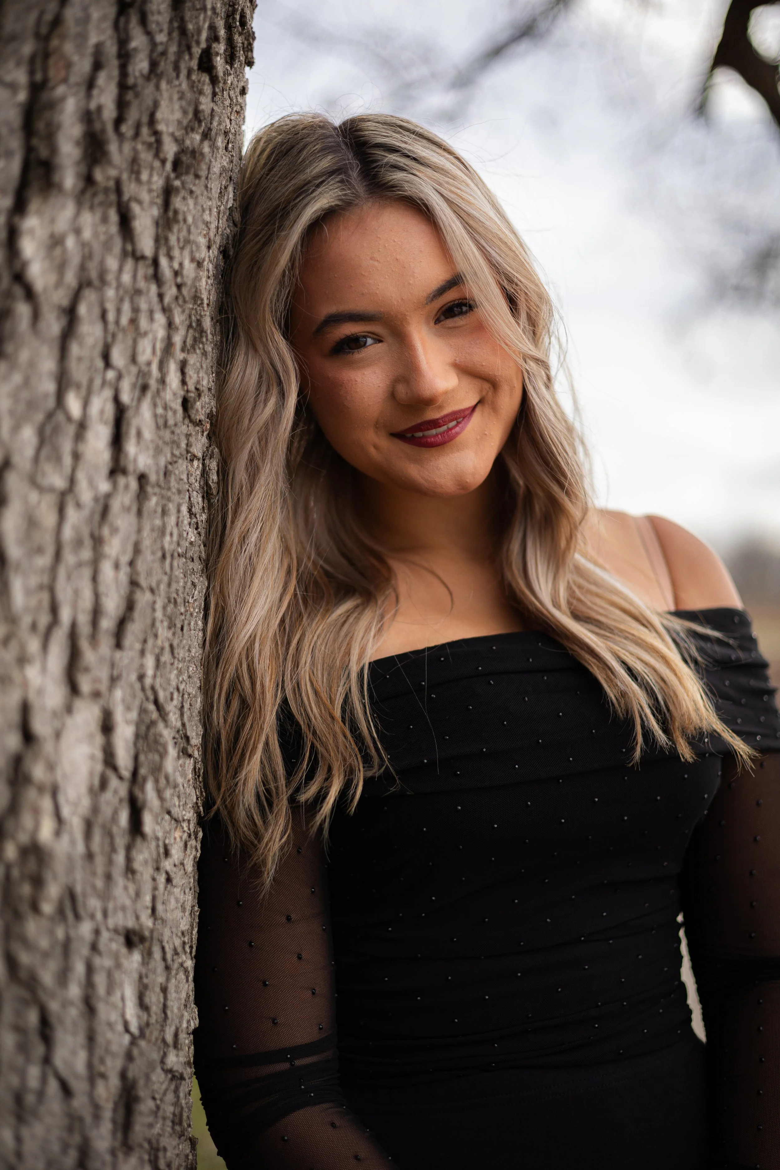 A young woman with blonde, wavy hair smiling while leaning against a tree outdoors, wearing a black dress with sheer, polka-dotted sleeves.