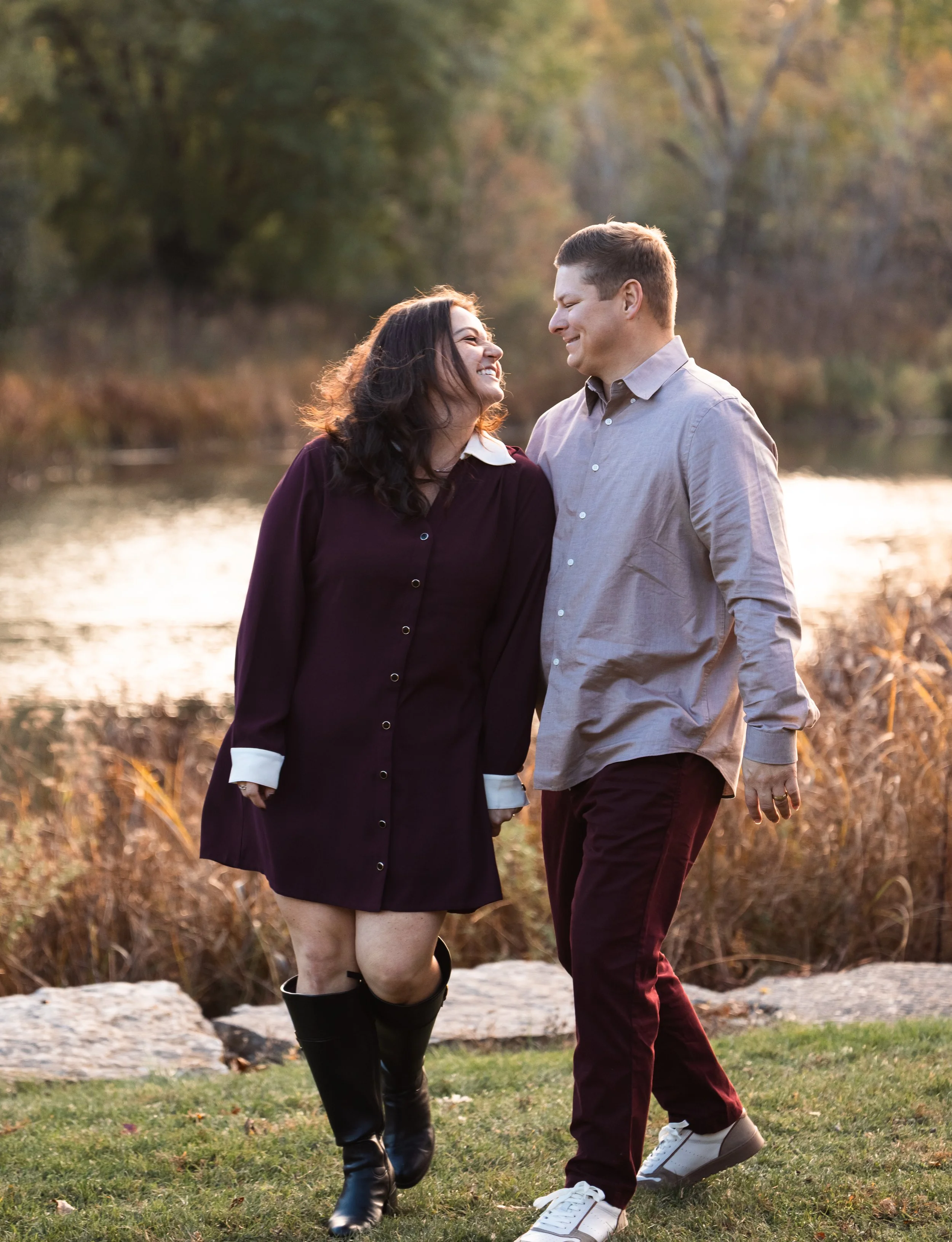 A smiling couple walking together outdoors near a lake or river during autumn, holding hands and enjoying each other's company. The woman is wearing a dark dress with white cuffs and black boots, and the man is wearing a light gray shirt with maroon 